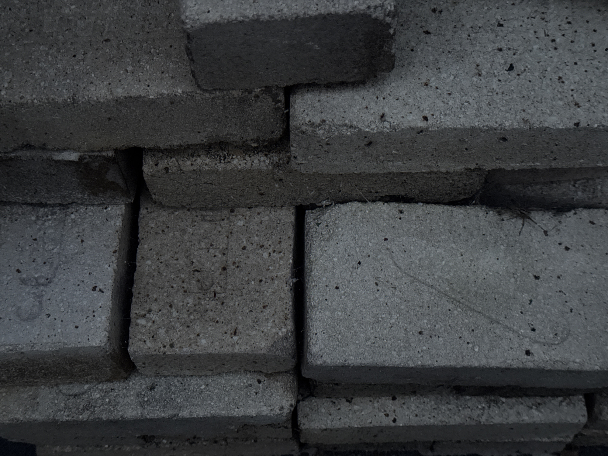 The image is a close-up, top-down view of a stack of grey concrete bricks. The bricks are rough in texture with visible aggregate and small dark flecks. They are stacked unevenly, with some bricks overlapping others. The lighting is dim, suggesting it might be overcast, dusk, or indoors with artificial lighting. There are no people visible in the image. The scene is devoid of any specific location cues beyond the material itself. The overall impression is one of raw, unrefined construction materials. The image does not contain any visible text. The location context of Kremenchuk, Ukraine, is not visually evident from the image content.