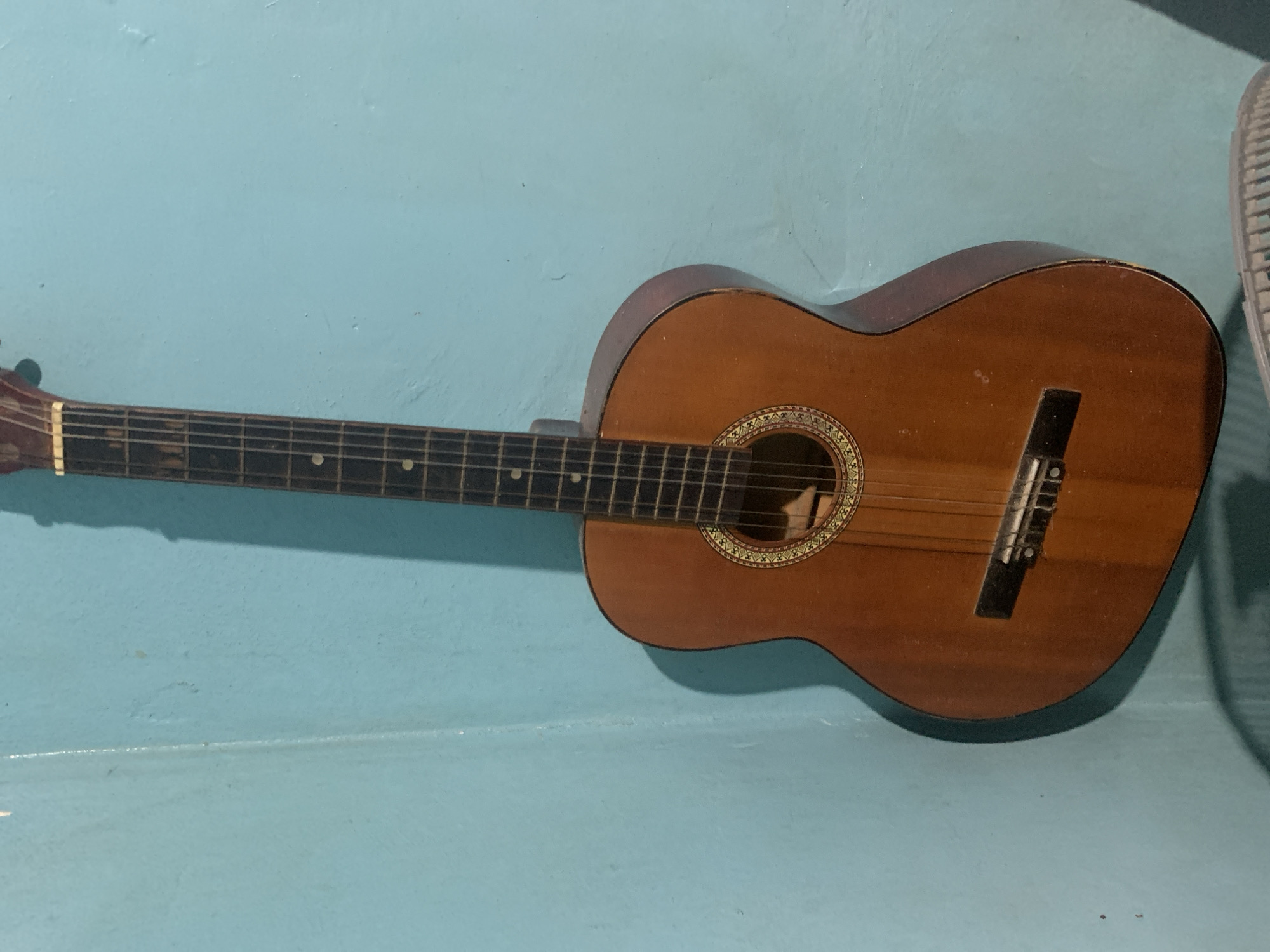 The image features a classical or acoustic guitar positioned horizontally against a light blue-green wall in what appears to be an indoor setting in Santa Ana, Venezuela. The guitar is made of warm brown wood and shows some minor wear or dust on its surface. A decorative rosette with intricate patterns encircles the soundhole. Six strings are visible, stretching from the tuning pegs (partially out of frame to the left) over the dark fretboard, which has white dot inlays, to the bridge on the guitar body. A segment of a light-colored fan grill is visible on the far right side of the frame, partially obscured. The lighting creates a clear shadow of the guitar on the wall, indicating a light source from above or the left. A horizontal line is visible across the lower portion of the light blue-green background, suggesting a corner or shelf. There are no people, explicit activities, or visible text in the image.