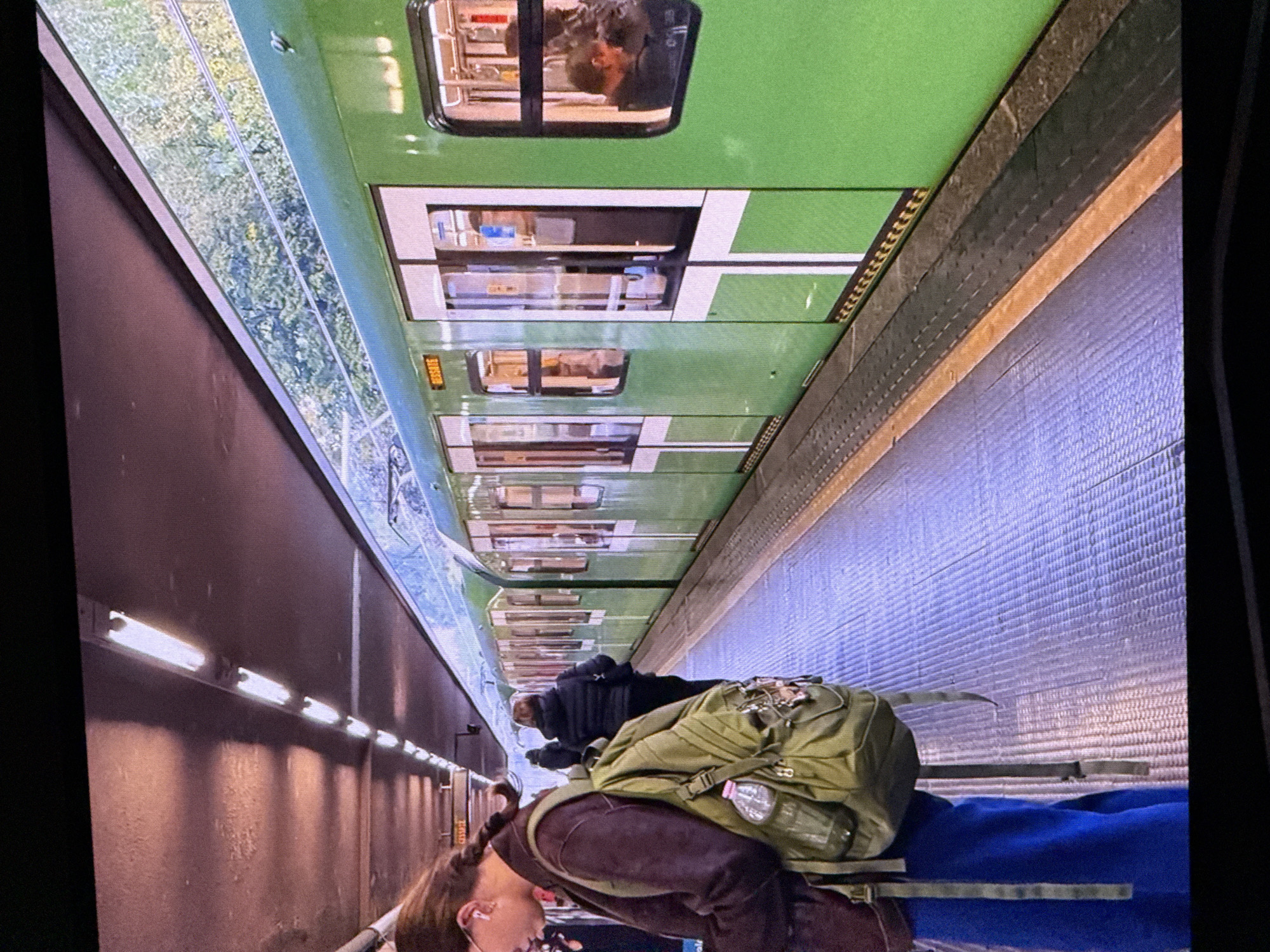 The image depicts a subway station platform in Milan, Italy, not Dubai. A person with braided hair wearing a dark jacket and a green backpack stands on the platform, facing away from the camera. The backpack has a water bottle attached. Further down the platform, two other figures are visible, one wearing a dark puffer jacket. A bright green subway train with white trim and multiple doors is stopped at the platform. The train's windows show passengers inside. The platform itself is tiled with a purple-blue pattern, bordered by yellow and black caution stripes and a darker grey area. Overhead, fluorescent lights illuminate the ceiling, casting shadows. The environment suggests an underground or covered station with greenery visible beyond the train.