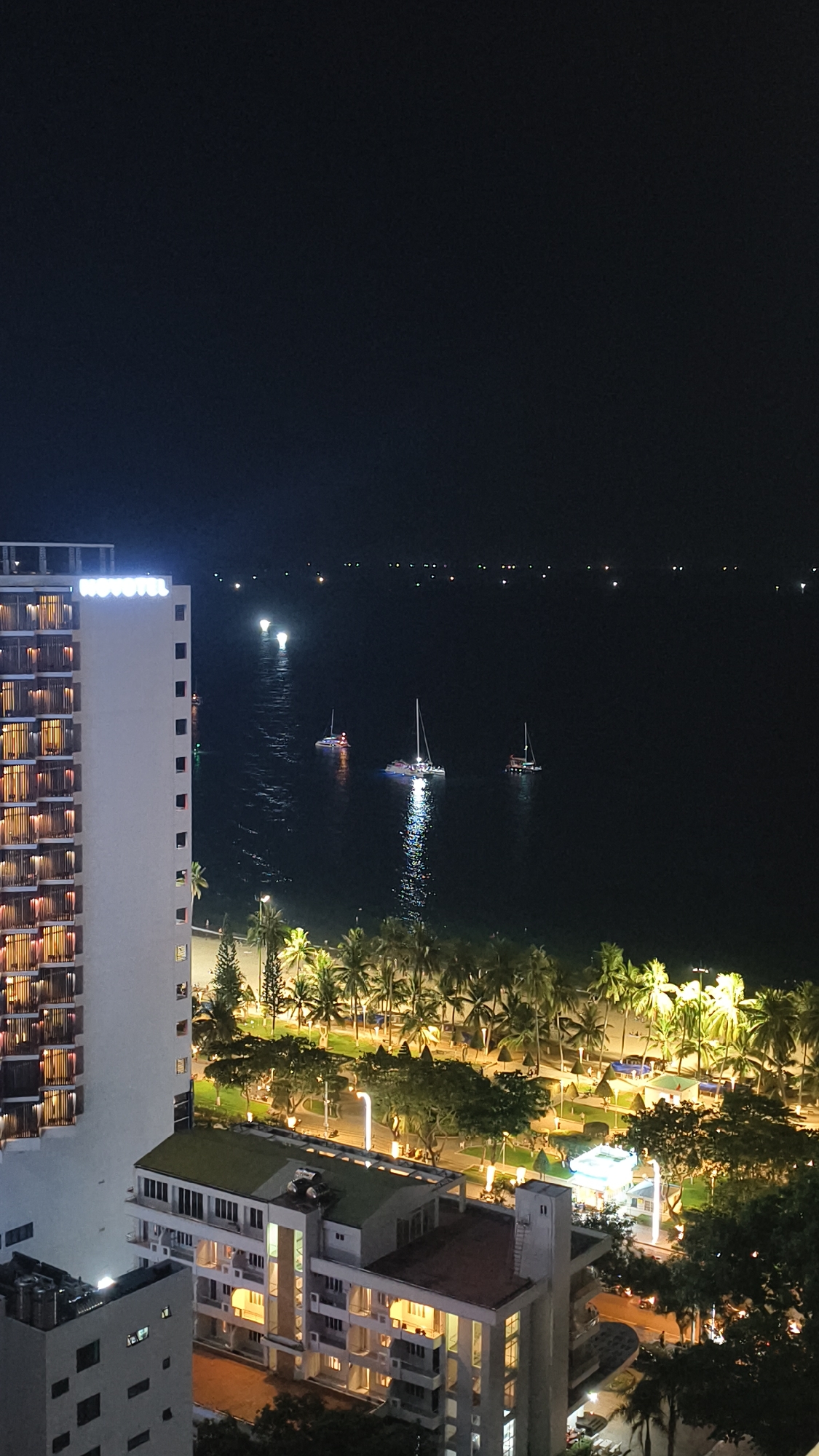 This is a nighttime aerial view of a coastal area in Nha Trang Ward, Vietnam. A tall, multi-story hotel, branded "NOVOTEL," stands on the left side of the frame, its windows illuminated. In the middle ground, a beach lined with palm trees is visible, with lights illuminating pathways and what appear to be beach umbrellas. Several catamarans and other boats are anchored or moving on the dark water of the sea. The distant horizon is dotted with small lights.