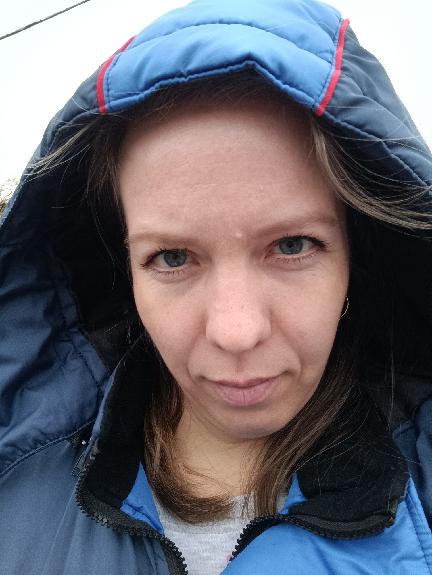 A woman with fair skin, light blue eyes, and shoulder-length light brown hair is looking directly at the camera with a somewhat unimpressed expression. She is wearing a blue quilted jacket with a hood, which is up and partially obscuring her hair. The hood has a red accent strip along its edge. The jacket is zipped up to her neck. The collar of a gray shirt is visible underneath the jacket. A small gold hoop earring is visible in her left ear. The background is a blurry, overcast sky. Based on the provided location information, the photo was taken in Chaikovsky, Russia. The specific location within the city is unknown.