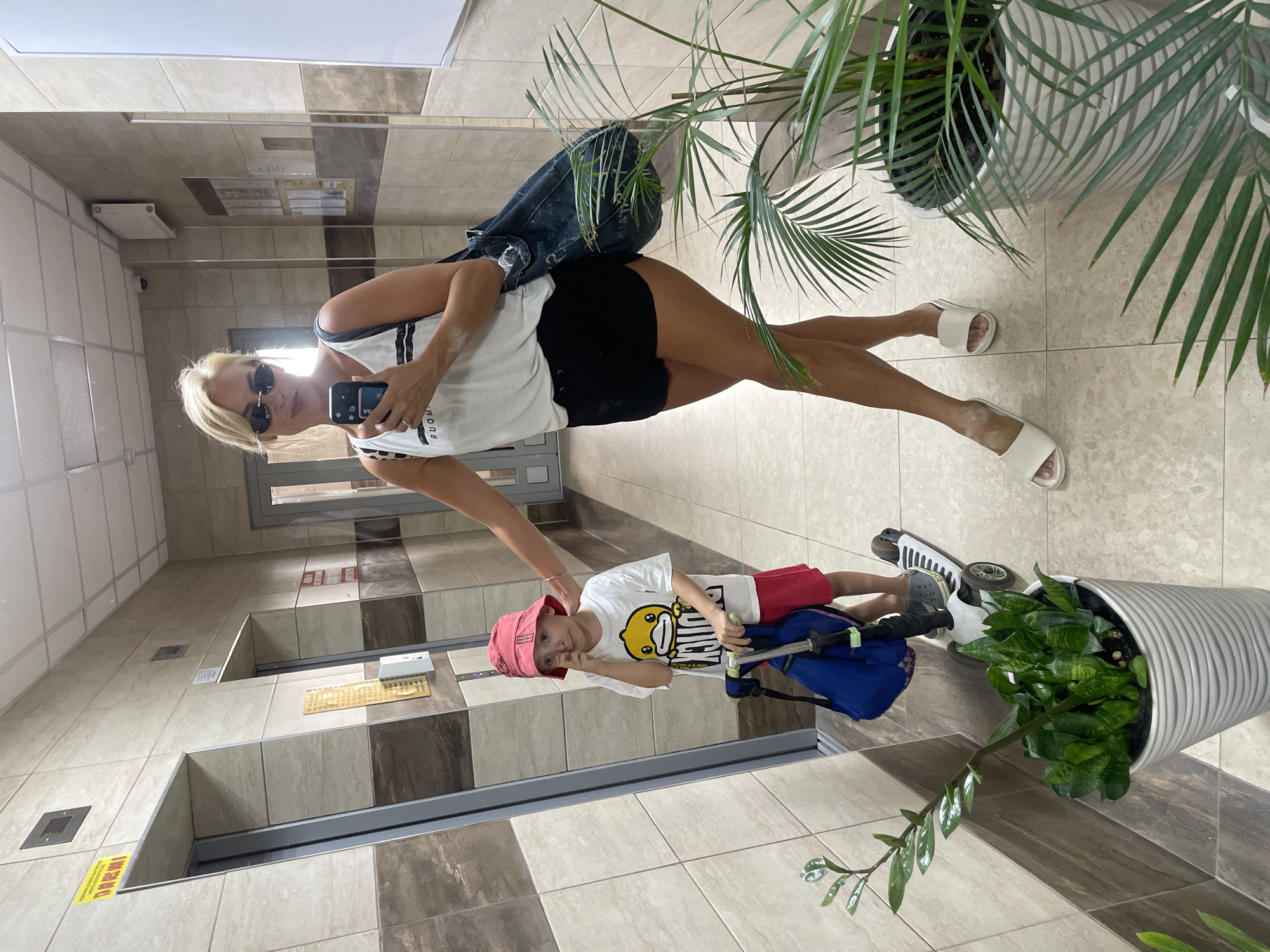 In an apartment building hallway in Novorossiysk, Russia, a woman with blonde hair and sunglasses takes a mirror selfie with her smartphone. She is wearing a white tank top, black shorts, and white slide sandals, and is carrying a denim tote bag. To her left, a young boy in a red bucket hat and white t-shirt stands next to a scooter. He is touching his nose with his finger. A potted plant is visible in the foreground. The hallway features tiled walls and two elevators.