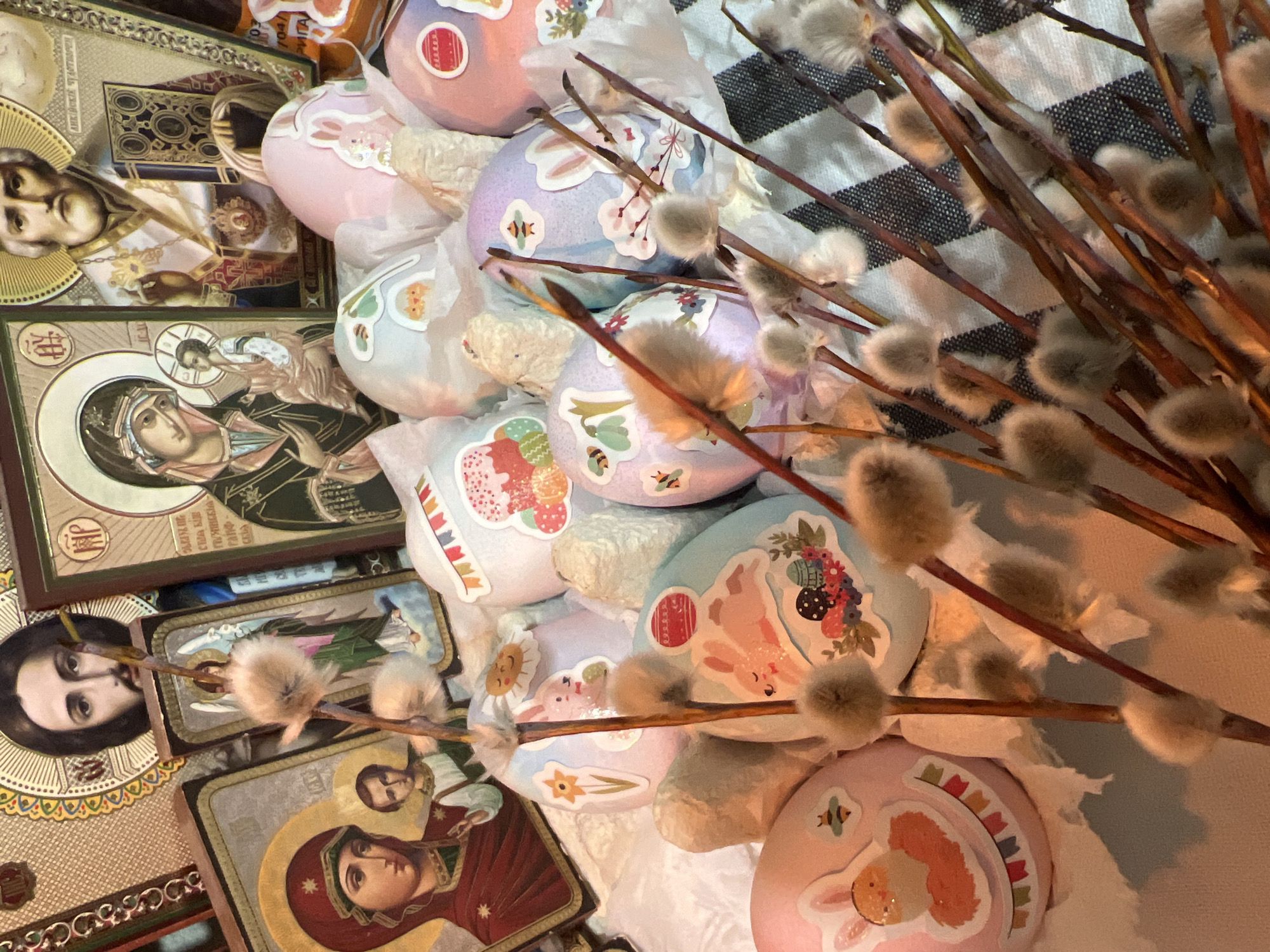 The image shows a still life scene composed of Orthodox icons, Easter eggs, and pussy willow branches. The icons, depicting religious figures, are arranged on the left side of the frame. The Easter eggs, decorated with stickers of rabbits and floral motifs, are piled in the center, interspersed with white fluffy material, likely cotton or artificial snow. Pussy willow branches are positioned on the right side, adding a natural element to the composition. The background appears to be a white surface with black stripes. It is assumed that the picture was taken in Kazan, Russia.