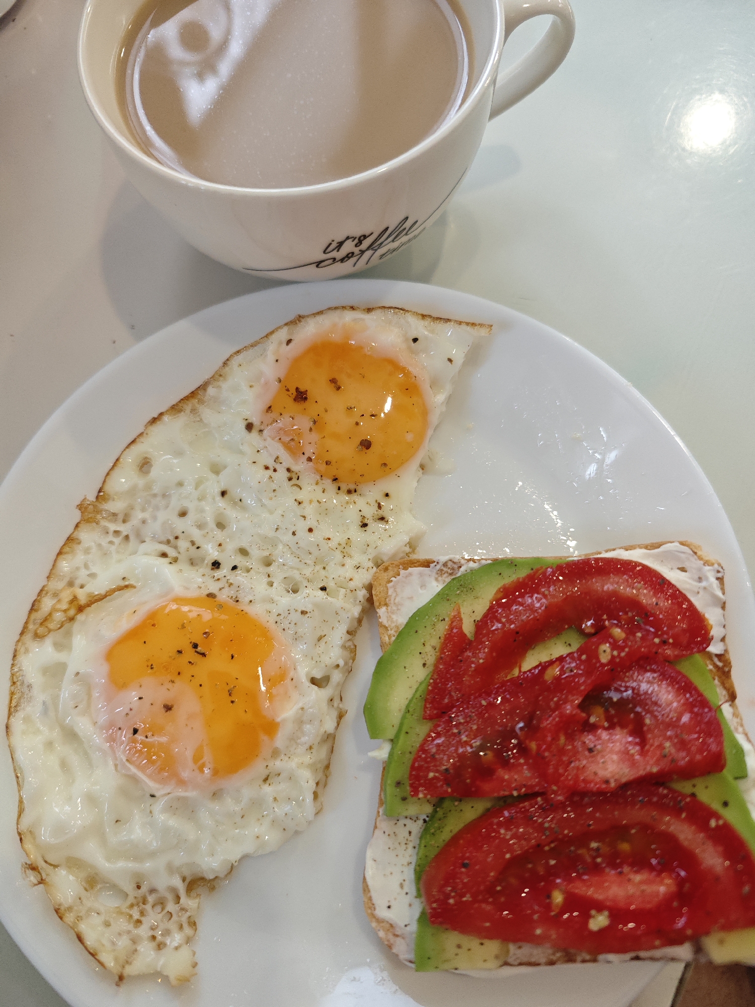 The image shows a breakfast meal on a white surface, likely a table. The meal consists of a white plate containing two fried eggs with visible yolks, seasoned with black pepper. Adjacent to the eggs is a slice of toast spread with a white creamy substance, topped with slices of avocado and tomato, also seasoned with black pepper. Above the plate is a white mug filled with a tan-colored beverage, possibly coffee or tea with milk. The mug has some black writing on it.
The location is not evident in this image. There is no information from the image to indicate that the image was taken in Chaikovsky, Russia.