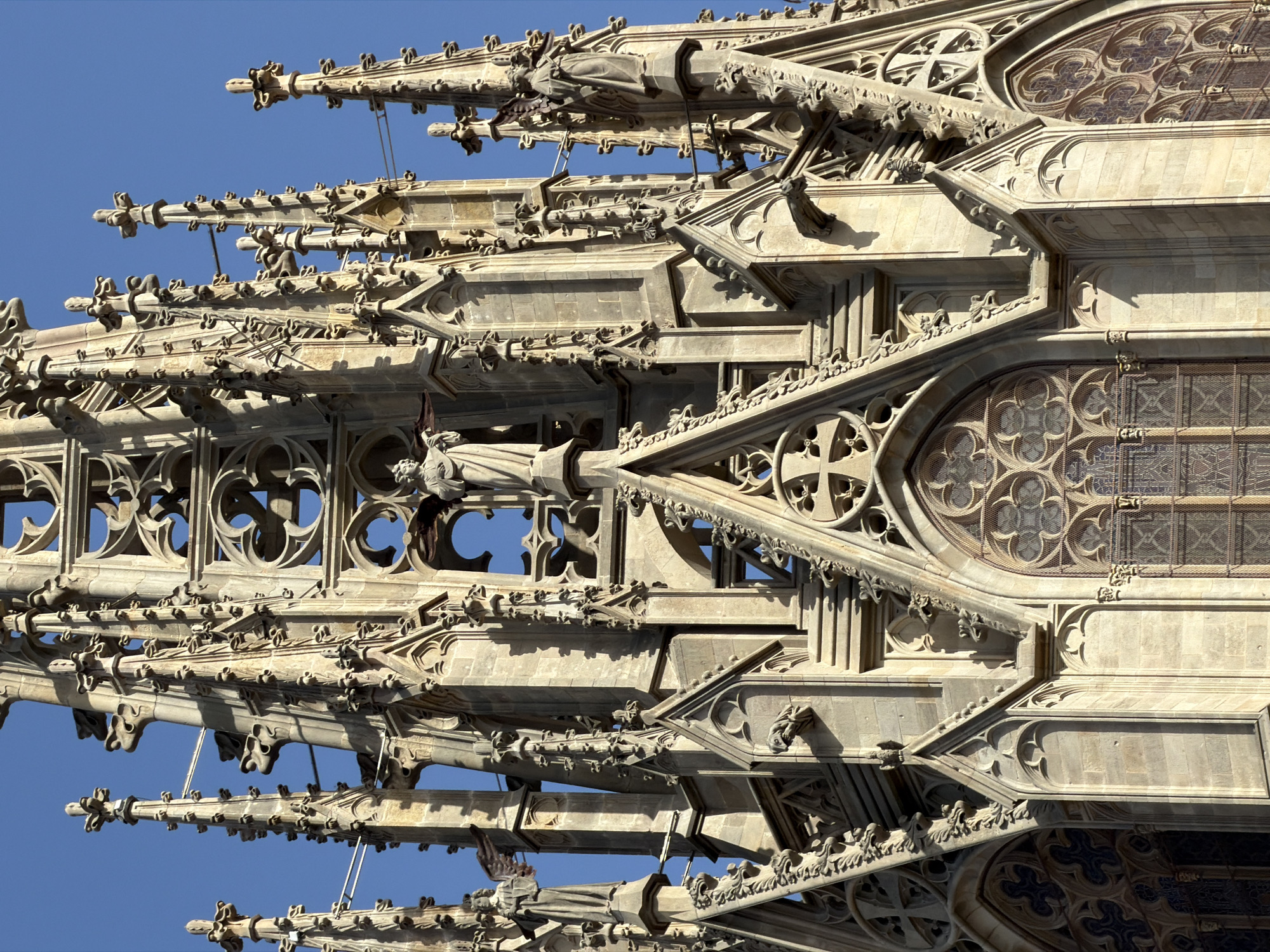 The image shows a close-up view of the spires and upper facade of the Barcelona Cathedral in Barcelona, Spain. The architecture is Gothic, characterized by intricate stonework, pointed arches, and delicate tracery. Several statues of figures, possibly saints or angels, are visible on the spires. Some of these statues appear to be winged. Gargoyles are also present, protruding from the stonework. The sky is clear and blue. The sunlight casts strong shadows, highlighting the textures and three-dimensional qualities of the stone carvings. The overall impression is one of immense detail and craftsmanship in religious architecture.