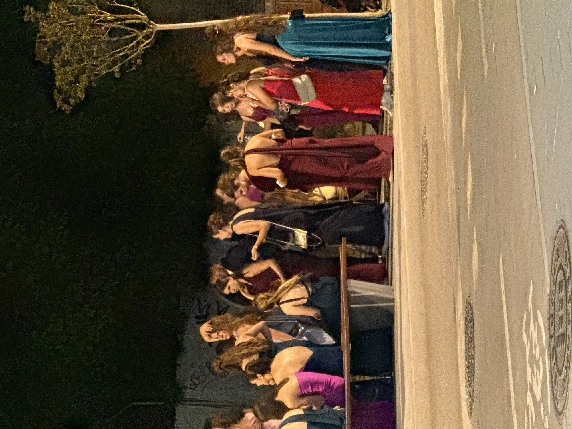 A group of people, predominantly young women, are gathered outdoors at night in what appears to be a public street in Blanes, Spain. They are dressed in formal attire, with many women wearing long, elegant dresses in various colors including red, burgundy, dark blue, and teal. Some are standing, others are seated on a low bench. The background features a dark, tree-covered area and a street with visible manhole covers and lane markings. The overall scene suggests a social gathering, possibly related to a formal event such as a prom or a wedding reception.