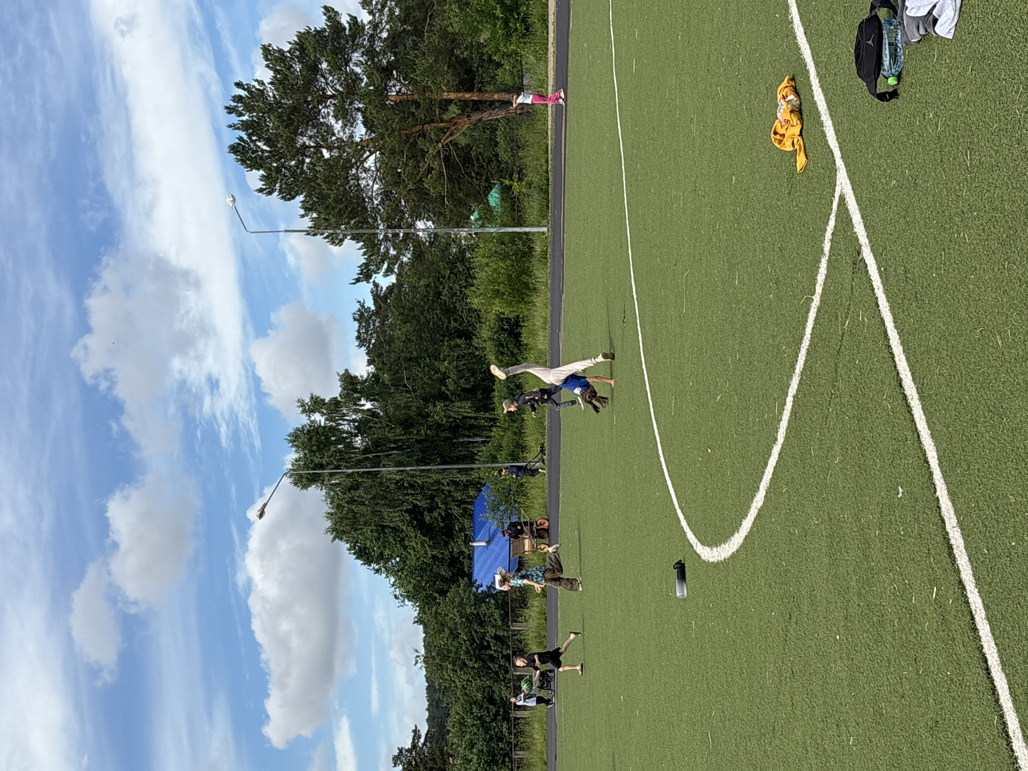 A group of children are engaged in various activities on an artificial turf field, possibly a sports complex, under a cloudy blue sky. In the foreground, a child is performing a cartwheel, with their legs in the air and body bent. To the left, another child in a dark t-shirt and shorts is running with a green object, possibly a ball, under their arm. Further back, a child in a patterned shirt and loose pants is also running. On the far left, two individuals are walking. In the background, there is a blue structure with a pointed roof, a vehicle with orange wheels, and a line of trees. A lamppost stands on the right side of the frame, and another is visible in the distance on the left. There are also several items scattered on the field, including a water bottle and a pile of clothes. The overall scene suggests a recreational or training session taking place outdoors. The location is identified as Chaikovsky, Russia.