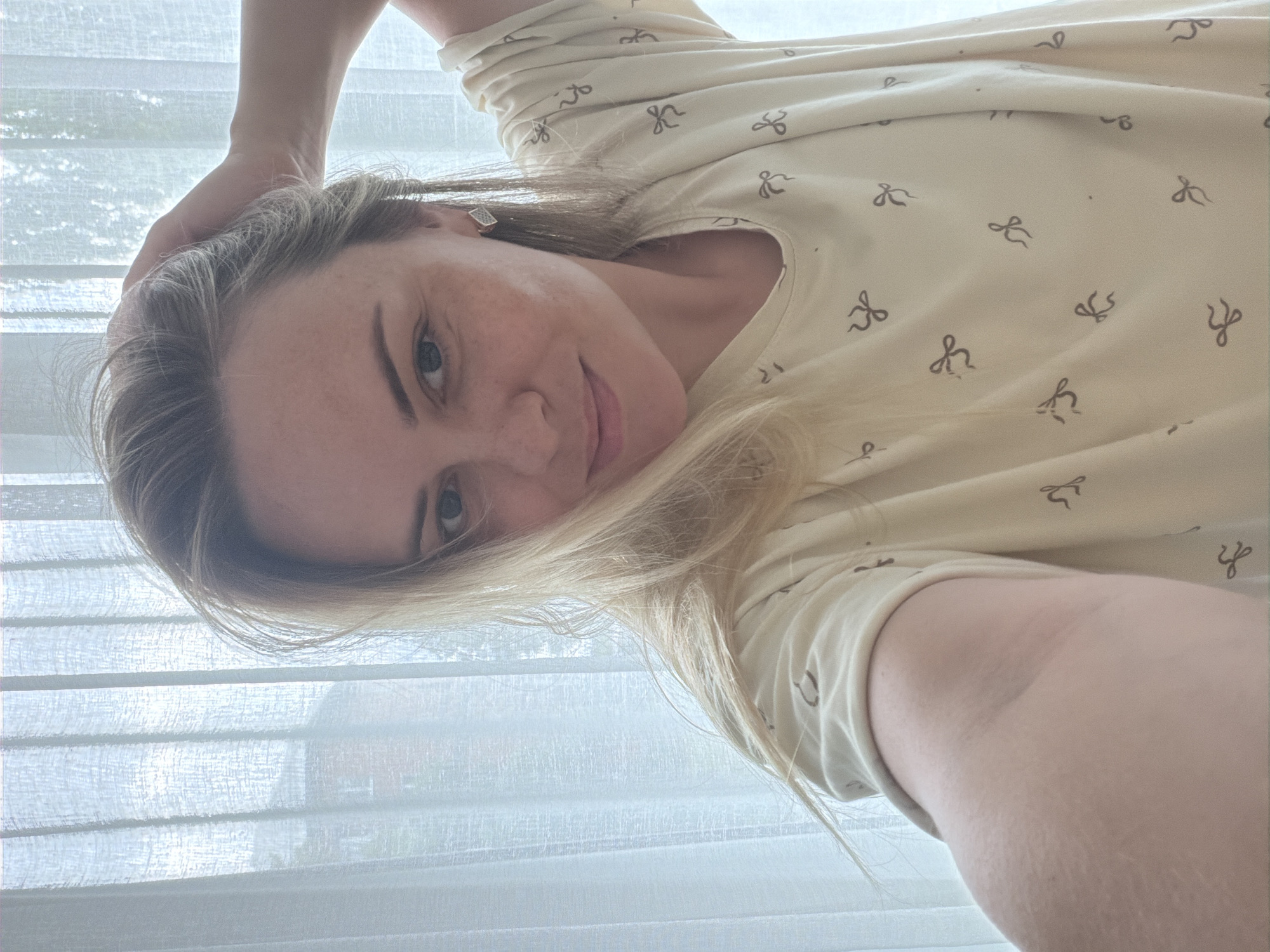 A woman with blonde hair and blue eyes is taking a selfie in front of a sheer white curtain. She is wearing a light-colored t-shirt with a pattern of small brown bows. Her left hand is raised and resting on her head, and she is smiling at the camera. The background shows a blurred view of buildings and trees outside. The lighting is soft and diffused, suggesting it is daytime.