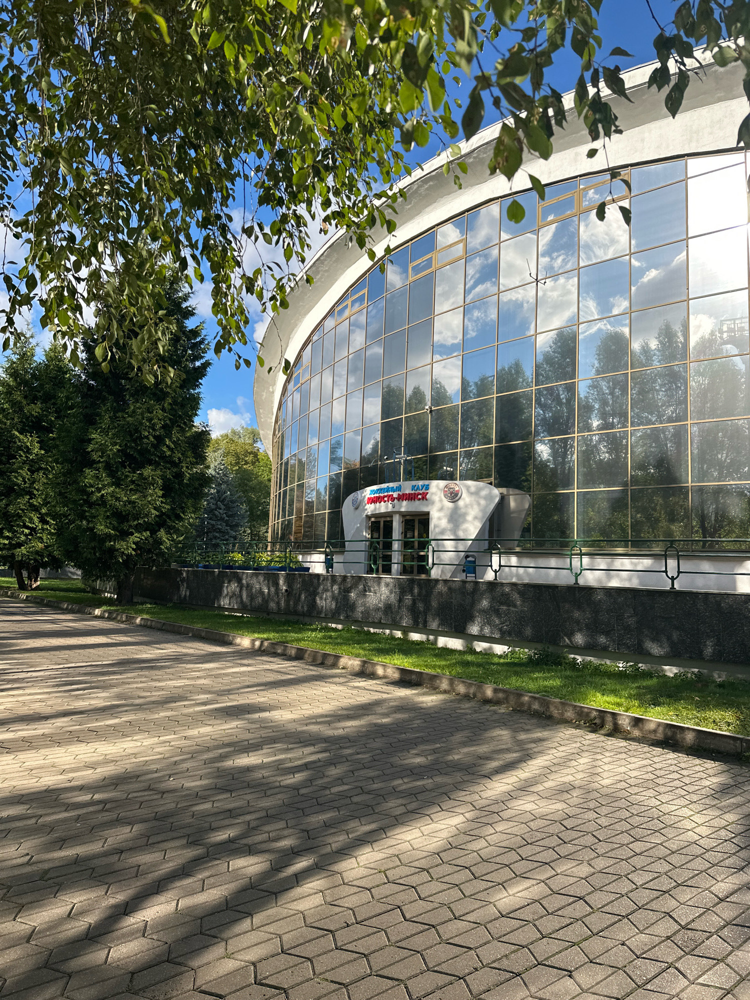 The image shows the exterior of a building with a large, curved glass facade. The glass reflects the blue sky with scattered clouds, and trees. A paved walkway with a herringbone pattern is in the foreground, casting long shadows from an unseen tree. The building has a sign above an entrance that reads "ХОКЕЙНЫЙ КЛУБ ЮНОСТЬ-МИНСК", indicating it is the Yunost-Minsk ice hockey club. The building appears to be located in Minsk, Belarus, given the signage. Green grass borders the walkway and the building. There are trees on the left side of the image, including a large evergreen tree.
