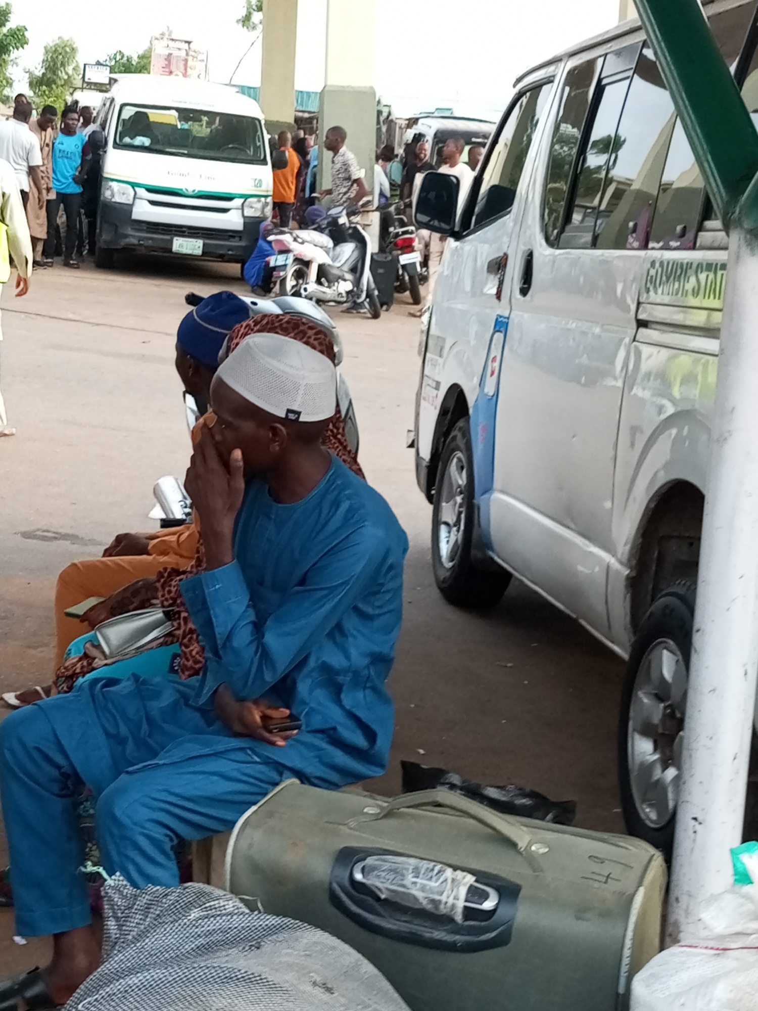 The image depicts a scene at a Gombe State transportation hub, likely a bus park, in Gombe, Nigeria. In the foreground, two men are seated, both wearing traditional attire. The man on the right, wearing a blue kaftan and a white cap, is seen with his hand to his face, possibly in thought or discomfort. He holds a mobile phone in his lap. Beside him, another man in an orange outfit is partially visible. A large, green cylindrical object, possibly a drum or a large bag, sits on the ground in front of them. Behind them, a white minibus with "GOMBE STATE" written on its side is parked, along with other vehicles and motorcycles. Several other people are standing or milling about in the background, suggesting a busy transport environment. The ground appears to be asphalt or concrete, typical of an urban setting. The overall atmosphere seems to be one of waiting and transit.