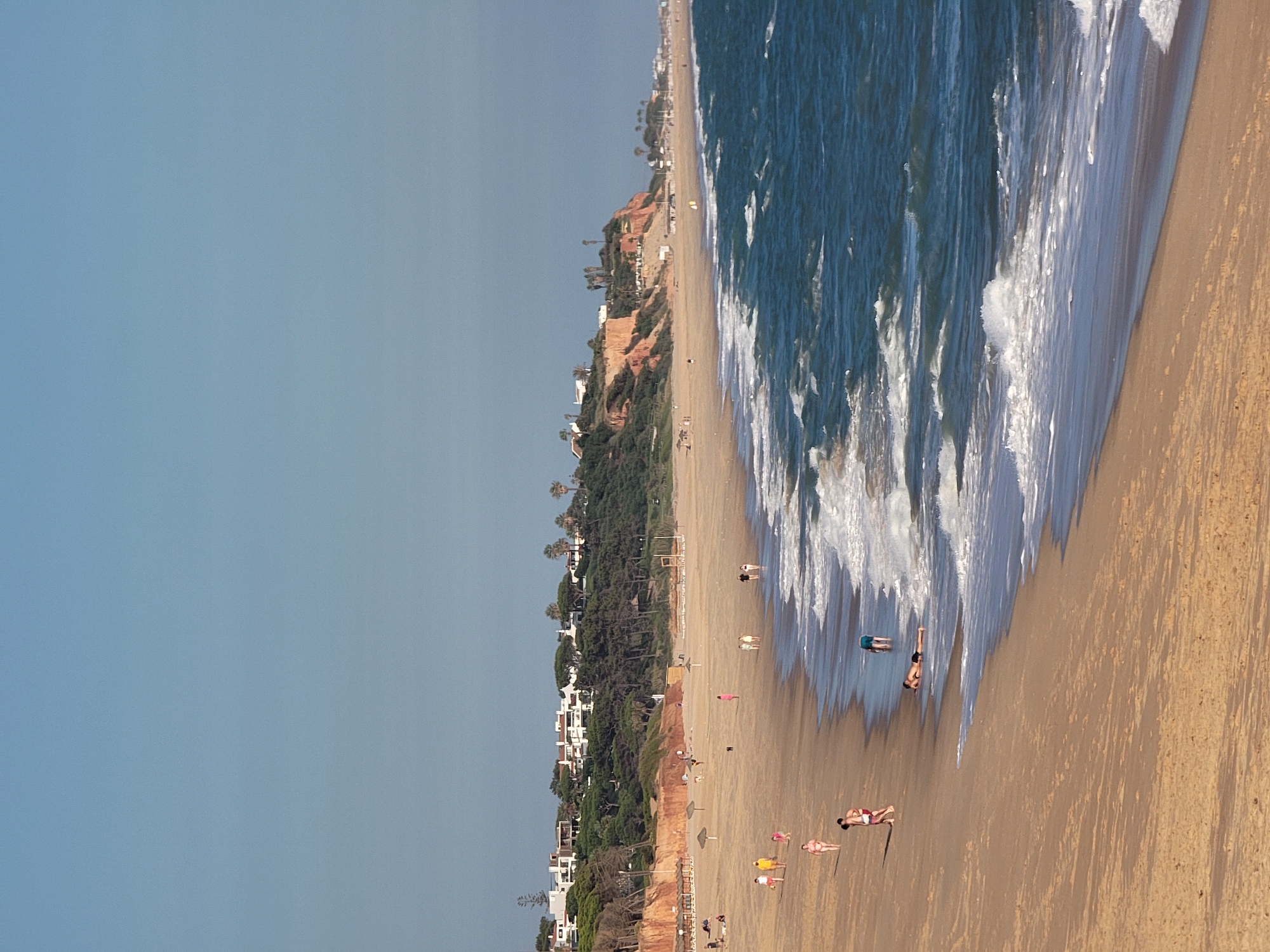 This is a medium-angle, eye-level, vertical shot of a wide, sandy beach with a line of residential buildings and dense green trees on a clifftop in the background. The sky is clear and pale blue. The sea is a deep blue with white-capped waves rolling towards the shore. Several people are visible on the beach, some walking, some lying down, and one person standing in the shallow water. The sand is smooth and light brown, with shadows cast by the people. In the distance, the beach extends further, and there are a few umbrellas and lounge chairs visible. The clifftop is covered in vegetation and dotted with white buildings, suggesting a resort or residential area. The overall impression is of a sunny, pleasant day at the beach in a developed coastal town. The location is likely the Praia da Falésia in Quarteira, Portugal, known for its distinctive red cliffs and expansive sandy beaches.