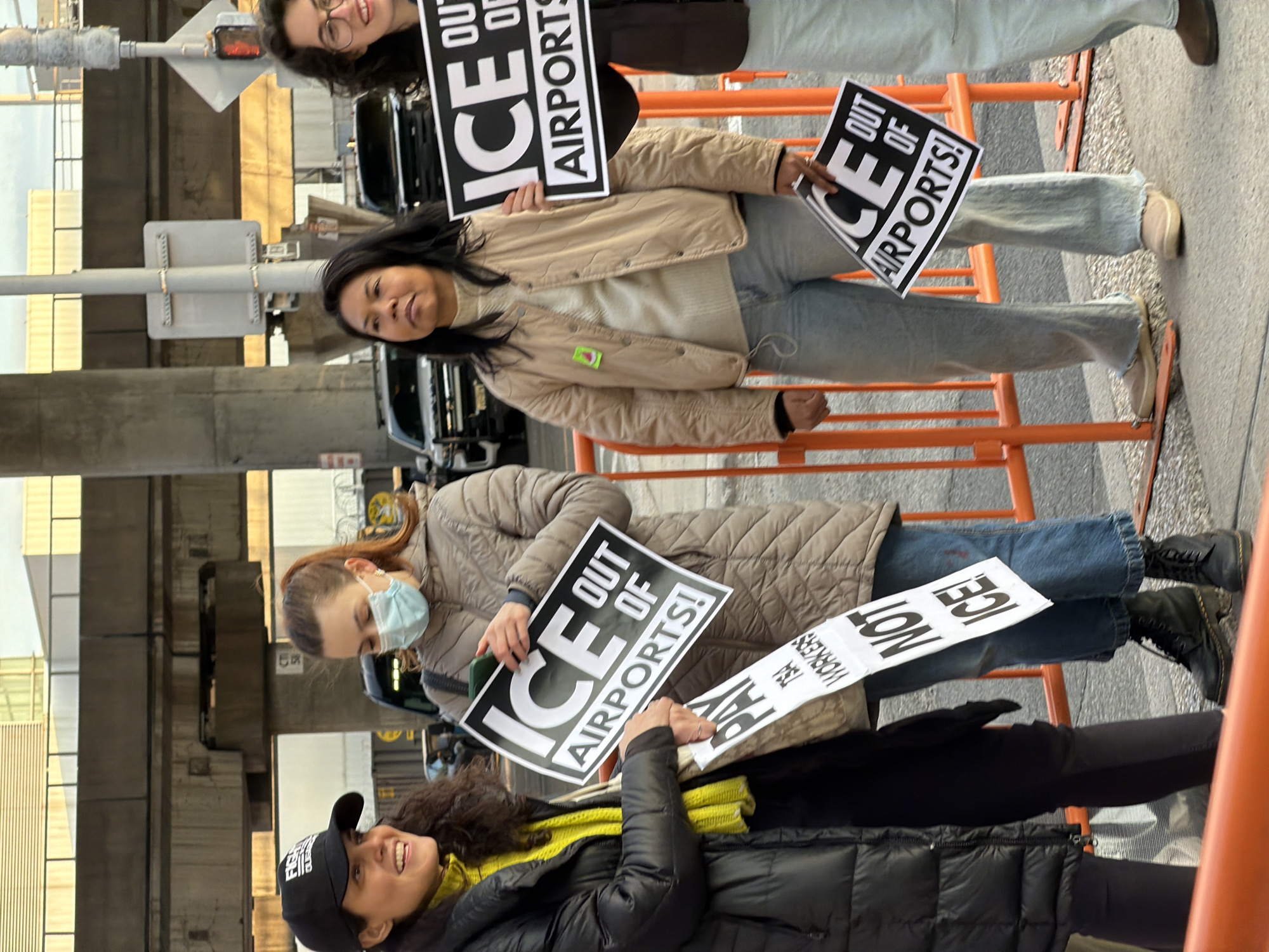 Four women participate in an outdoor demonstration at an airport facility in New York, United States of America. They stand behind orange plastic safety barriers on a paved surface.
The leftmost woman, wearing a black baseball cap, yellow turtleneck, and black puffer jacket, smiles towards the right. She holds two signs: one reads "PAY TSA WORKERS" and the other, partially visible below it, "NOT ICE!".
Next to her, a woman with brown hair in a ponytail wears a light blue surgical mask, beige quilted jacket, blue jeans, and black lace-up boots. She holds a sign with both hands that states "ICE OUT OF AIRPORTS!" in large black letters on a white background. She appears to be looking at a green smartphone held in her left hand.
The third woman from the left, with dark hair, wears a light beige quilted jacket with a green pin, a light beige turtleneck, light blue jeans, and light brown slip-on shoes. She holds a smaller "ICE OUT OF AIRPORTS!" sign in her right hand, positioned lower than the other signs, and looks slightly upward.
The rightmost woman, partially visible, has dark curly hair and wears glasses, a brown jacket, and light blue pants. She also holds an "ICE OUT OF AIRPORTS!" sign and smiles towards the left.
In the background, an elevated concrete overpass structure is visible, supported by a pillar marked "CTI 5B". Dark-colored vehicles are parked or moving on a roadway behind the barriers. A tall light-colored pole and a partial traffic signal are also present. The environment appears to be an urban area with infrastructure consistent with an airport.