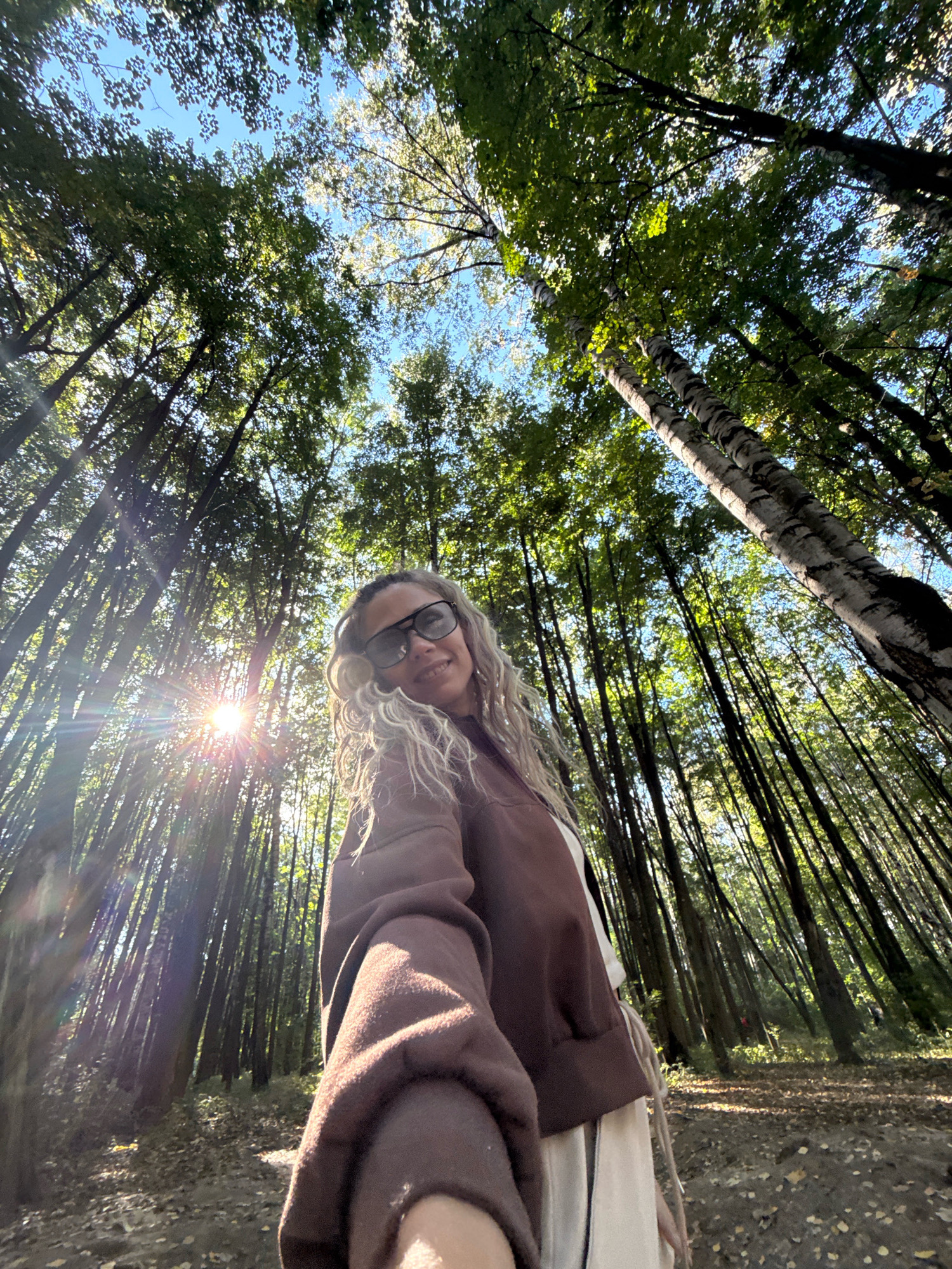 A woman with curly blonde hair and sunglasses is taking a selfie in a forest. She is wearing a brown hoodie over a white shirt and off-white pants. The sun is shining through the trees, creating lens flare. The forest floor is covered with fallen leaves, and the trees are tall and slender. The scene appears to be in the city of Rusanovo, Russia.