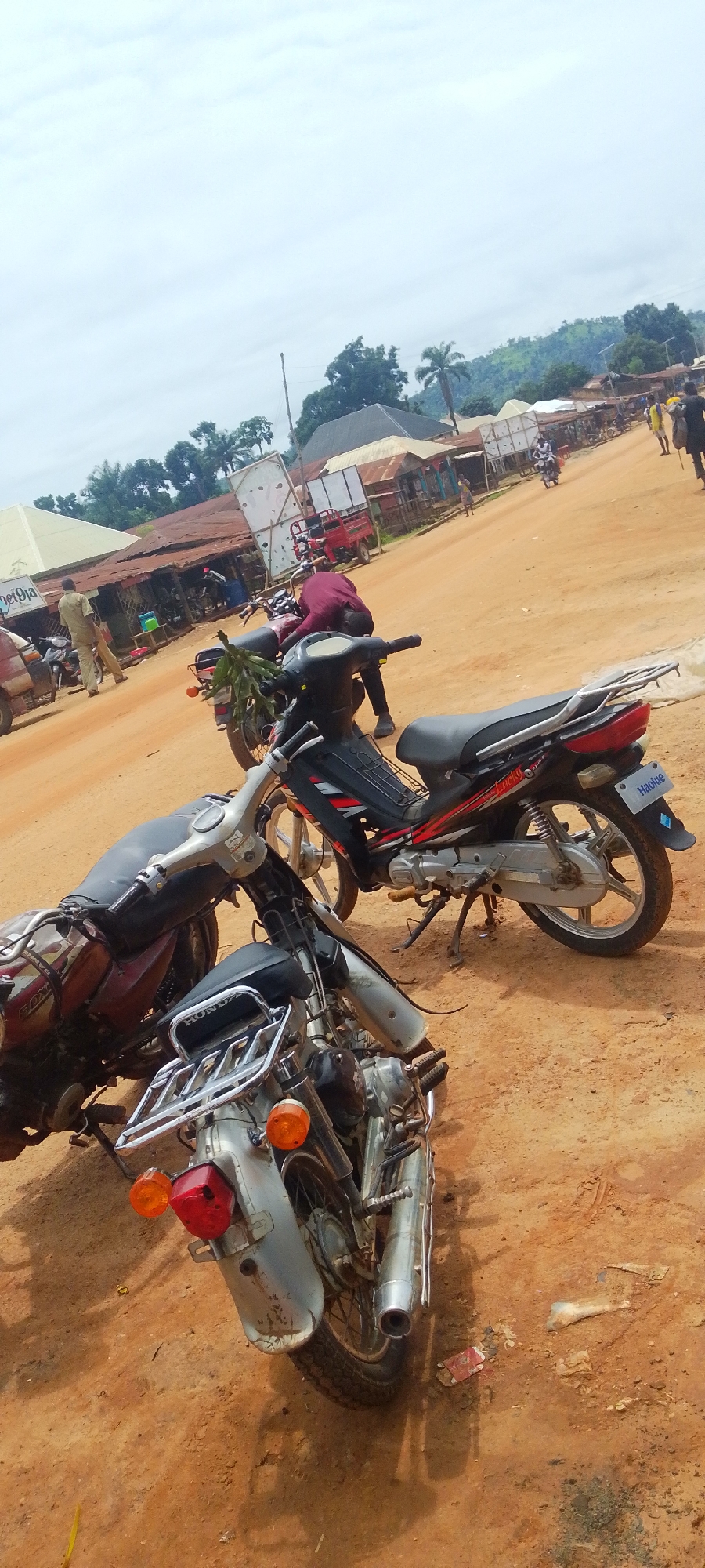 The image depicts a street scene in Baissa, Nigeria. The foreground is dominated by two parked motorcycles, one a silver and black model with a chrome luggage rack and the other a darker, possibly maroon, motorcycle partly obscured. In the middle ground, a person in a dark maroon shirt is bending over a motorcycle, seemingly working on it. To the left of this person, another motorcycle, black with red accents, is parked. Further in the background, a dirt road stretches into the distance, lined with modest buildings, some appearing to be shops with corrugated metal roofs. Several people are visible along the road, some walking and others near the buildings. One person in a light-colored shirt stands near a building on the left. Another person in a yellow shirt is further down the road, carrying a bag. A red cart, possibly a type of delivery vehicle, is parked near one of the buildings. The overall scene suggests a busy, possibly market day, in a rural or semi-urban Nigerian setting. The sky is overcast, indicating a cloudy day. The terrain appears somewhat hilly in the far background, with lush green vegetation. Visible text includes "Lucky" and "Haolue" on the black and red motorcycle.