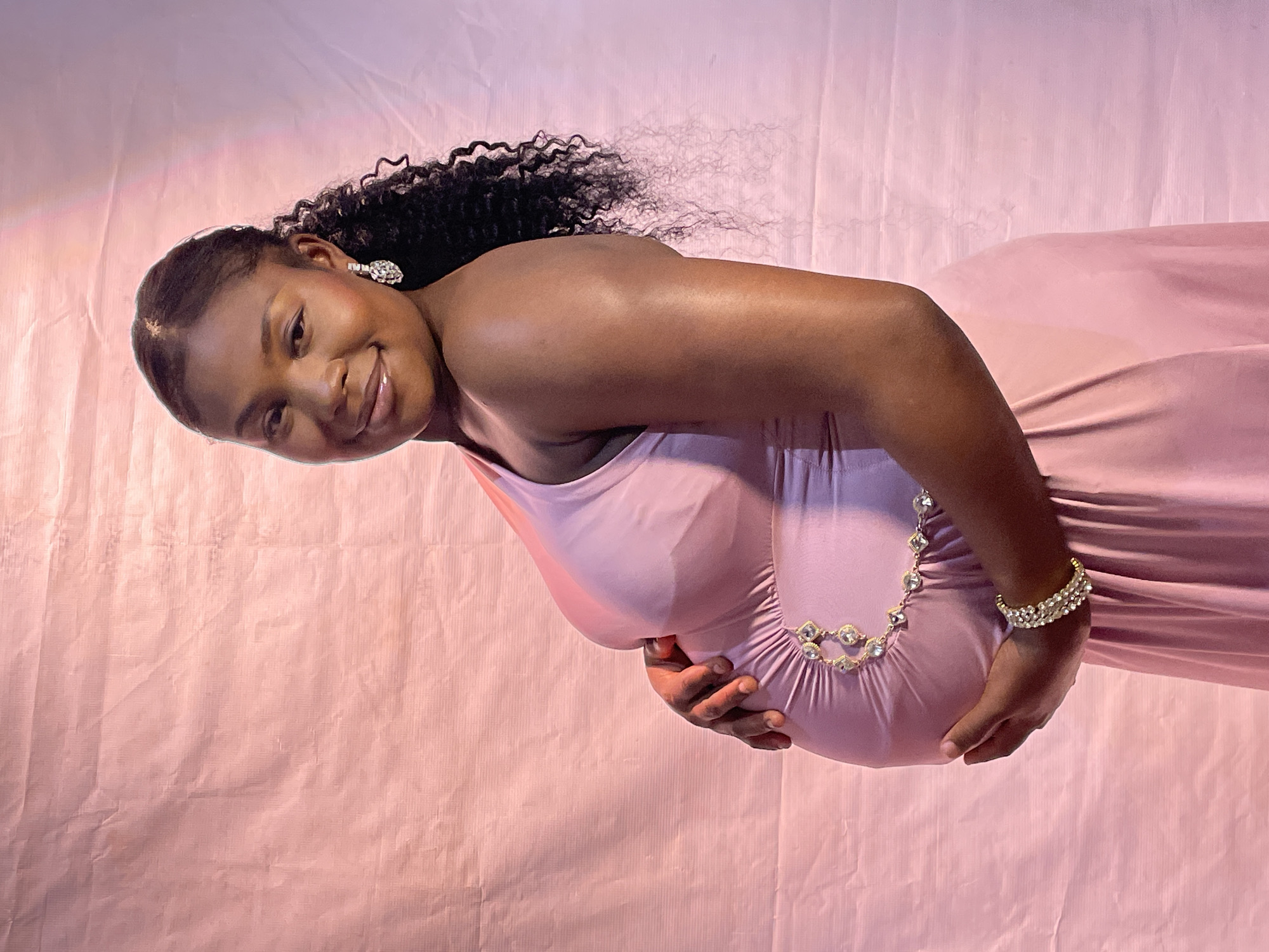 A pregnant woman with dark skin and curly black hair is turned sideways in a photo against a pink backdrop. She is wearing a pink dress, a bracelet on her right wrist, and earrings. She is holding her stomach. The location is Jos, Nigeria.