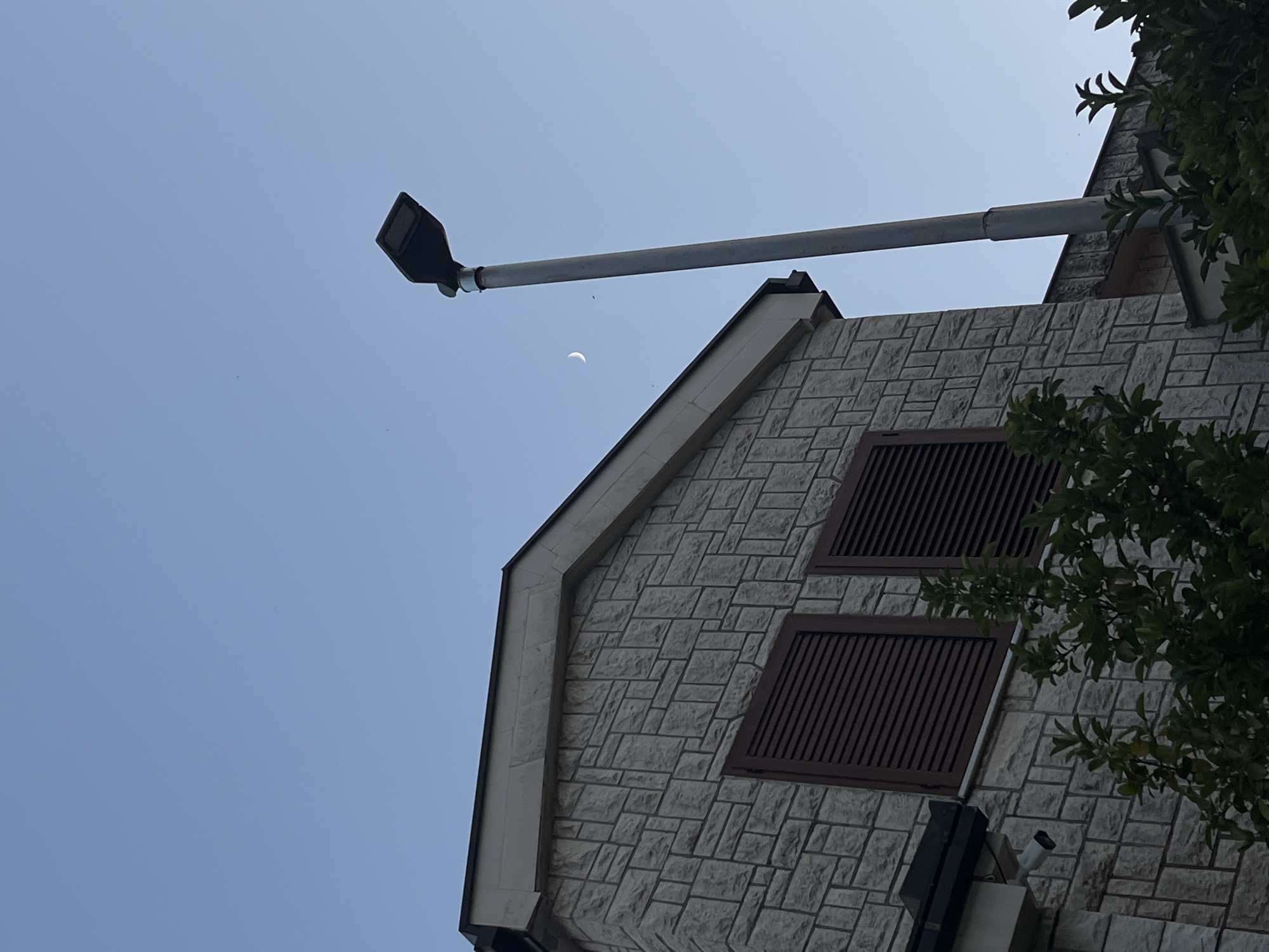 This is a low-angle, medium-shot, eye-level photograph of a stone-clad building and a street lamp against a clear blue sky in Budva, Montenegro. The building's facade is constructed from light-colored, rectangular stones laid in an offset pattern. Two windows with dark brown louvered shutters are visible on the building. A security camera is mounted to the left of the windows, near the roofline. In the foreground, dark green foliage obscures the lower portion of the building. A tall, gray street lamp stands to the right of the building, its head tilted slightly. A crescent moon is visible in the sky between the building and the street lamp, along with a few small, dark specks that may be birds or distant objects. The overall lighting suggests either early morning or late afternoon.