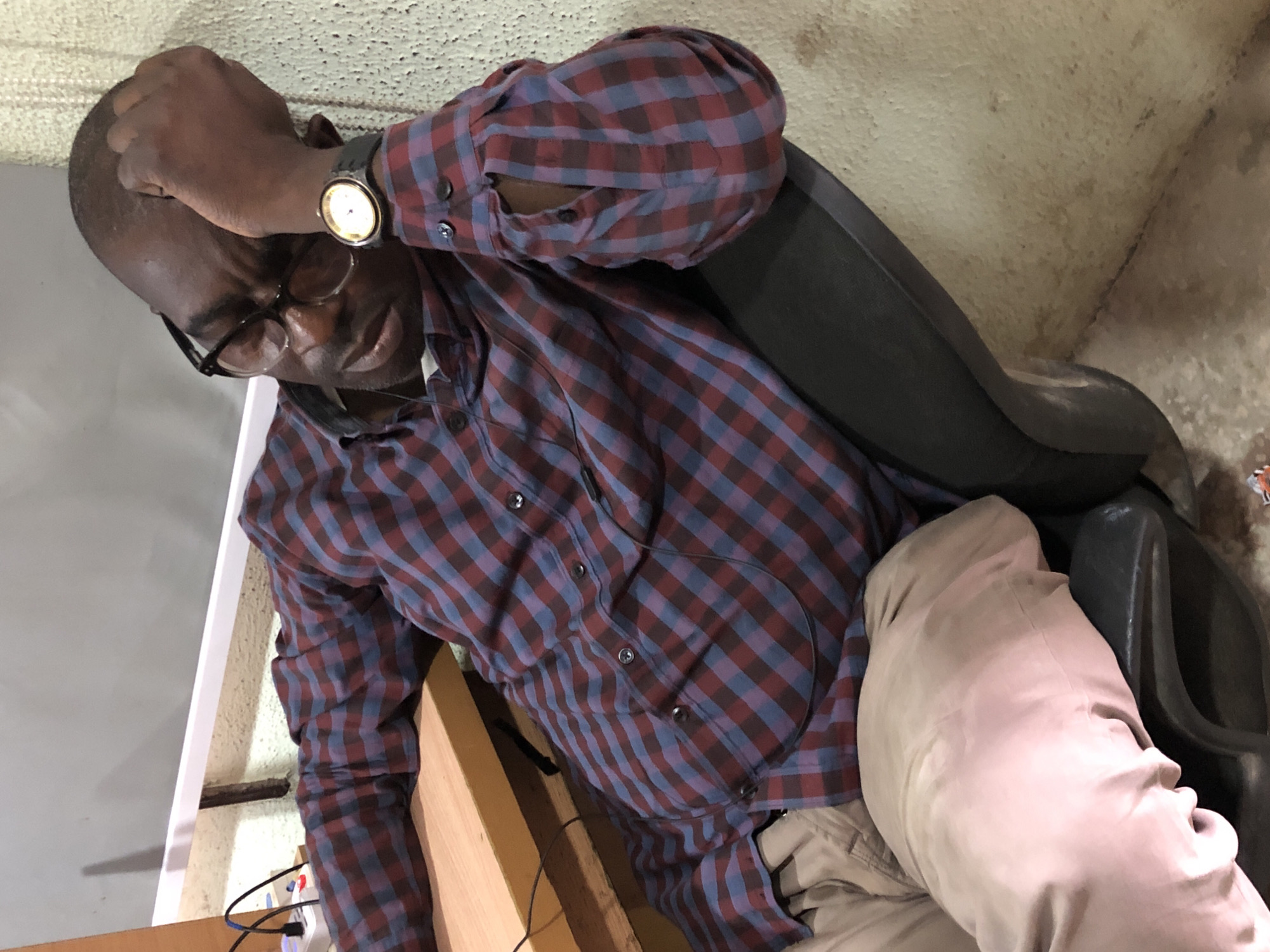A dark-skinned man with short hair and glasses is leaning back in a black office chair. He is wearing a red and blue plaid long-sleeved shirt and tan pants. His right hand is resting on his forehead, and his eyes are closed. A watch with a gold face and black band is visible on his left wrist. In the background, there is a desk with a power strip and some wires, and a textured wall. The scene appears to be in an office in Ikeja, Nigeria.