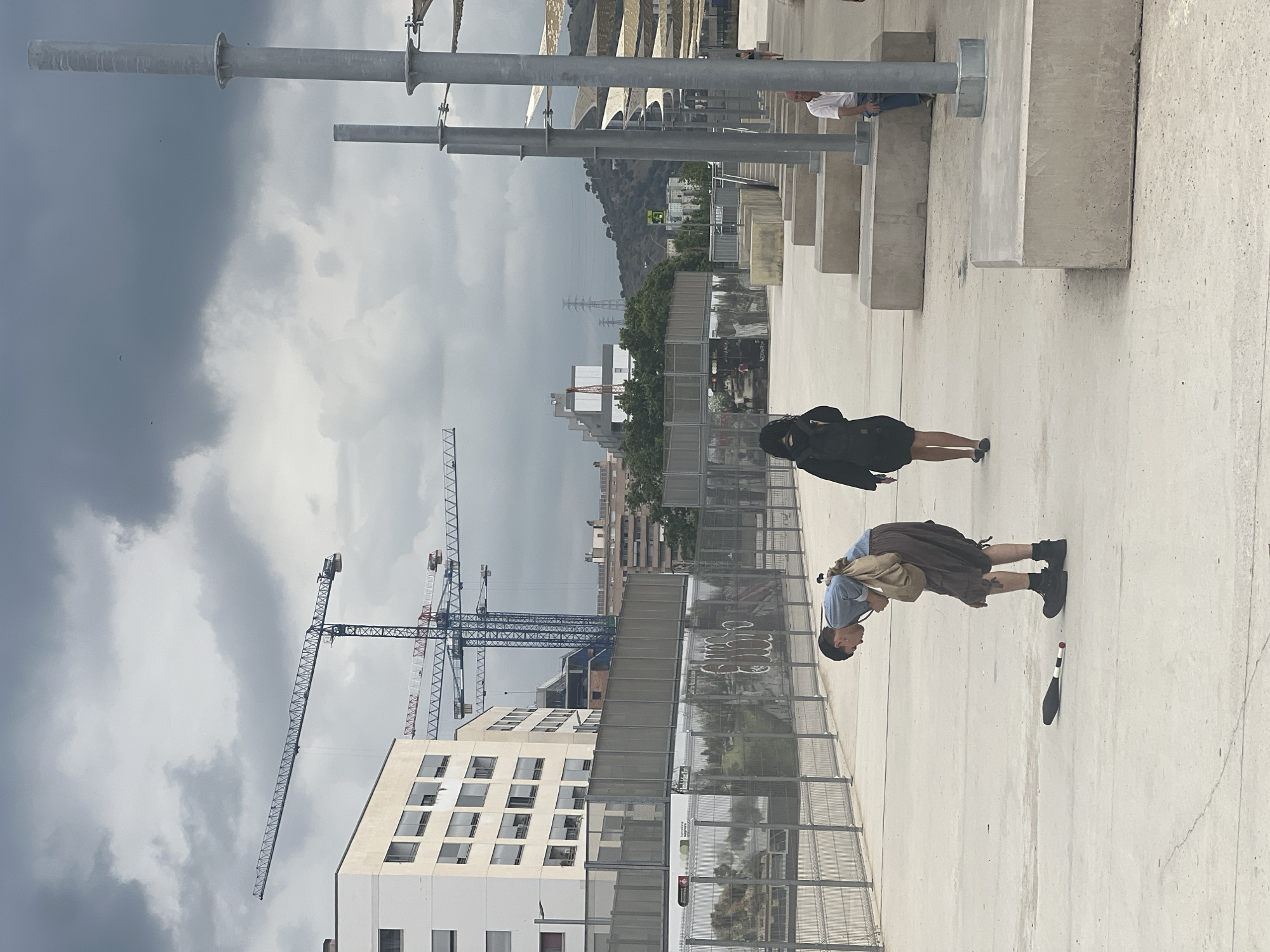 A medium shot captures a person in the foreground, dressed in a blue shirt and brown shorts, bending down to pick up a juggling pin lying on a light-colored concrete ground. They are carrying a beige bag over their shoulder. To the left, a chain-link fence encloses an area with posters and graffiti. In the background, several tall construction cranes dominate the skyline against a cloudy sky, with a modern white building on the left and a distant hillside with power lines. To the right, a woman in a black dress walks away from the camera, and further back, a man in a white shirt and blue jeans is seated on concrete blocks. The overall scene appears to be an outdoor urban area under construction, possibly a public space or park adjacent to a construction site, in Barcelona, Spain.