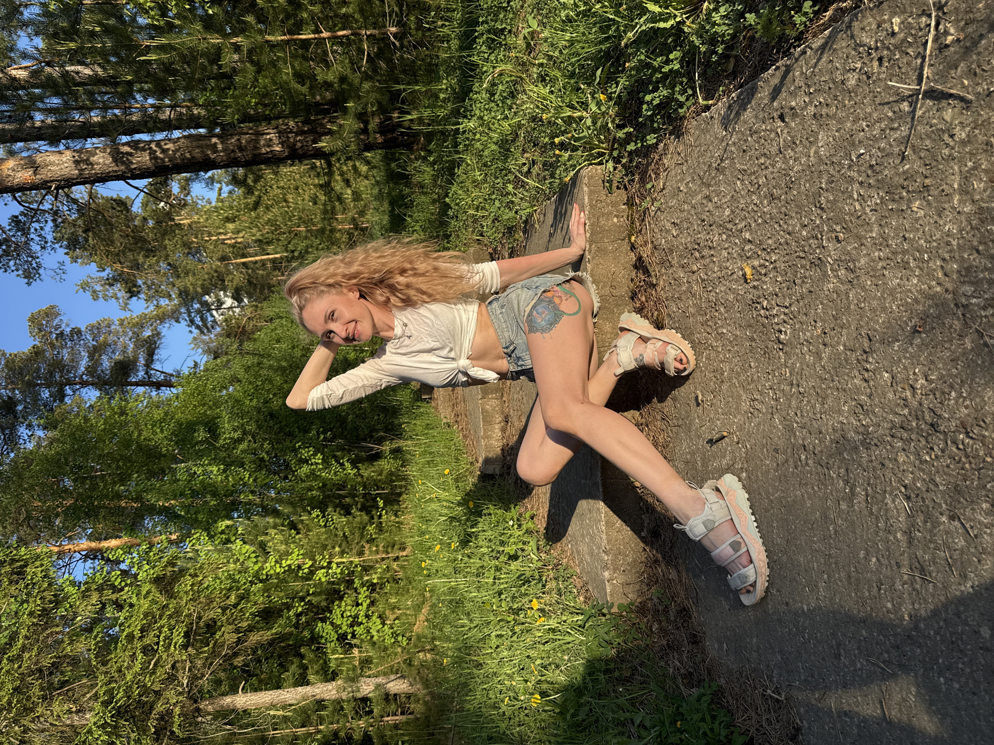 A woman with long blonde hair is posing on a concrete structure next to a grassy area in what appears to be a natural setting. She is wearing a white knotted shirt and denim shorts, with sandals on her feet. Her left hand is on the concrete, while her right hand is resting on her head. The background includes trees and vegetation, suggesting a park or wooded area. The location is Noviy, Russia.