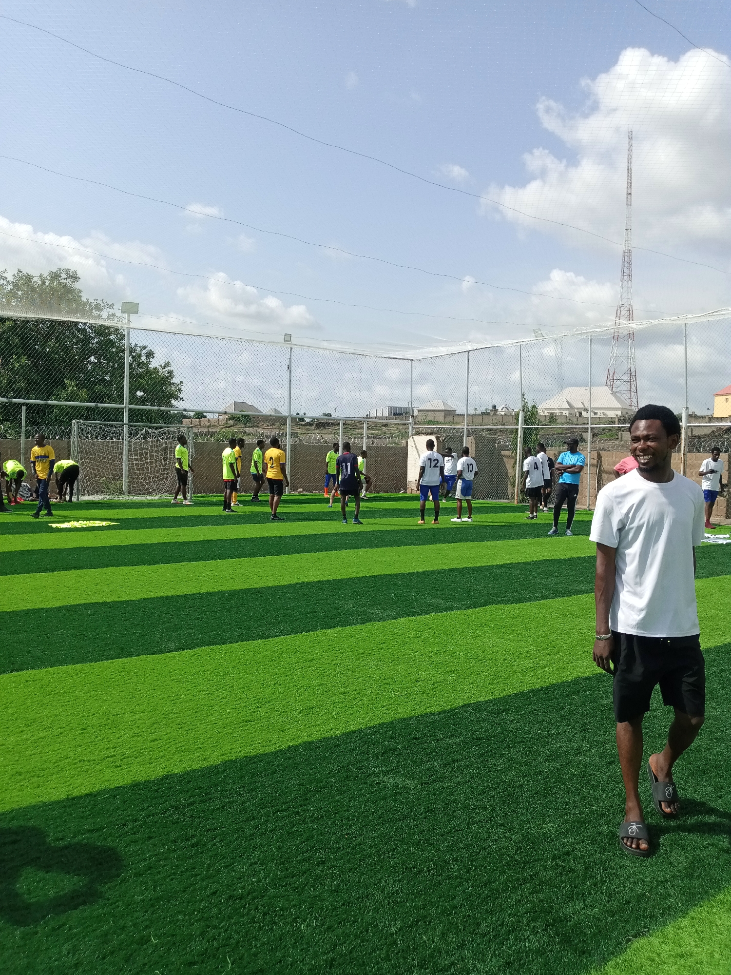 In Jimeta, Nigeria, an outdoor artificial turf soccer field is the setting for a group of young men engaged in soccer activities. The field, characterized by its vibrant green artificial grass striped with darker green lines, is enclosed by a chain-link fence and netting. In the foreground, a young man in a white t-shirt and black shorts stands facing the camera with a wide smile, his sandals visible. In the background, several other individuals are scattered across the field, some in yellow shirts and others in white shirts with numbers. Some appear to be playing soccer, while others are observing or resting. The sky above is a light blue with scattered clouds, and a red and white radio tower stands tall in the distance. The overall scene depicts a casual sports gathering on a bright, sunny day.