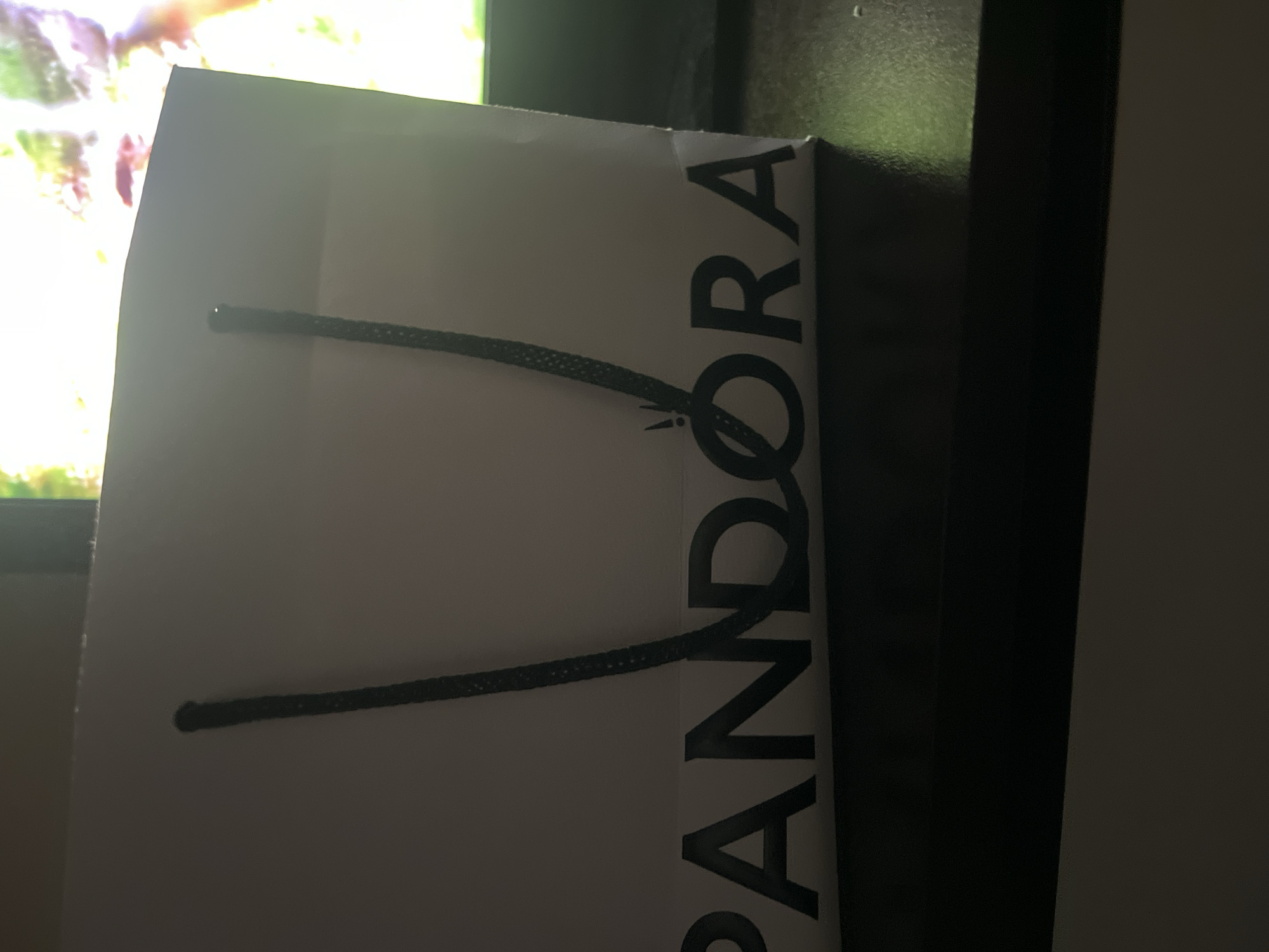 A white Pandora shopping bag with black handles is centered in the frame. The Pandora logo is printed in black on the lower portion of the bag. The bag is placed on a dark surface, possibly a table, in front of a brightly lit screen, which appears to be a television displaying a colorful image. The overall lighting is dim, with the screen providing the primary light source. There are no people or animals visible in the image. The background is out of focus due to the shallow depth of field. The image does not provide any specific geographical information or context that would allow for its placement in Petrevene, Bulgaria.