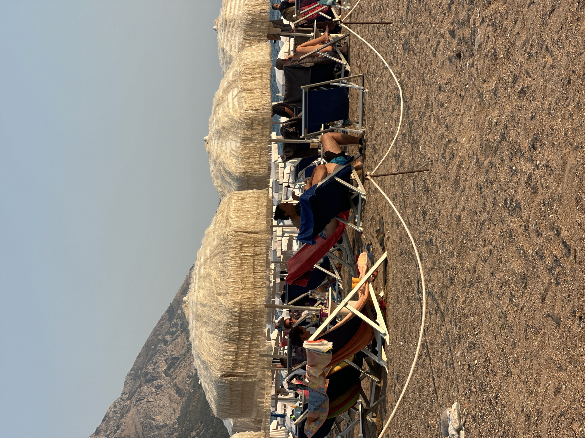 The image depicts a beach scene with several people relaxing on lounge chairs under large, thatched umbrellas. In the foreground, the sandy beach is visible, with a rope barrier running horizontally across the frame. Several lounge chairs are set up on the sand, some occupied by individuals. In the background, more umbrellas and lounge chairs can be seen, extending further down the beach. The sea is visible in the distance, and a mountainous landscape forms the backdrop under a clear sky. The lighting suggests it is late afternoon or early evening. The scene appears to be in Boreti, Montenegro, a coastal town known for its beaches and scenic views.