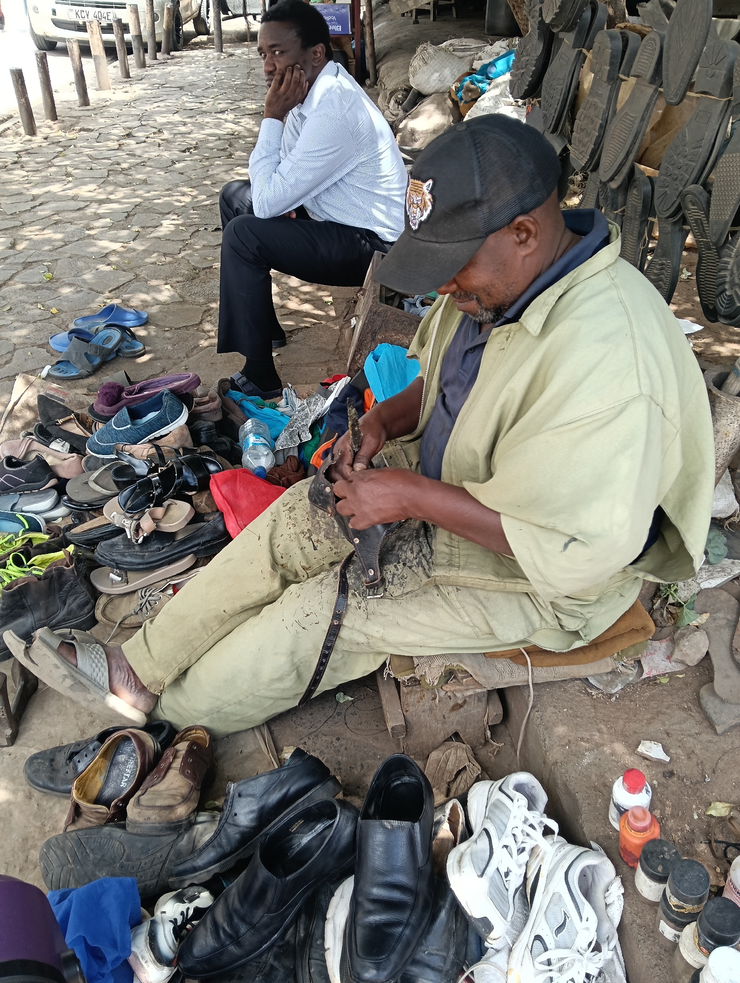 The scene shows a cobbler mending a shoe while seated on a wooden stool in an outdoor setting. The cobbler, wearing a black baseball cap with a tiger emblem, a light green jacket, and tan pants, is focused on his work. He is surrounded by a variety of shoes and sandals in various states of disrepair. To his right, there are bottles and containers, possibly containing glue or other shoe repair materials.
In the background, a man in a white button-down shirt and dark pants sits with his head resting in his hand, possibly a customer waiting.
The ground around the cobbler is dusty, and the backdrop includes a building, stone-paved sidewalks, and posts. There are hanging shoe soles visible behind the cobbler. The license plate of a vehicle indicates this scene is in Nairobi, Kenya.