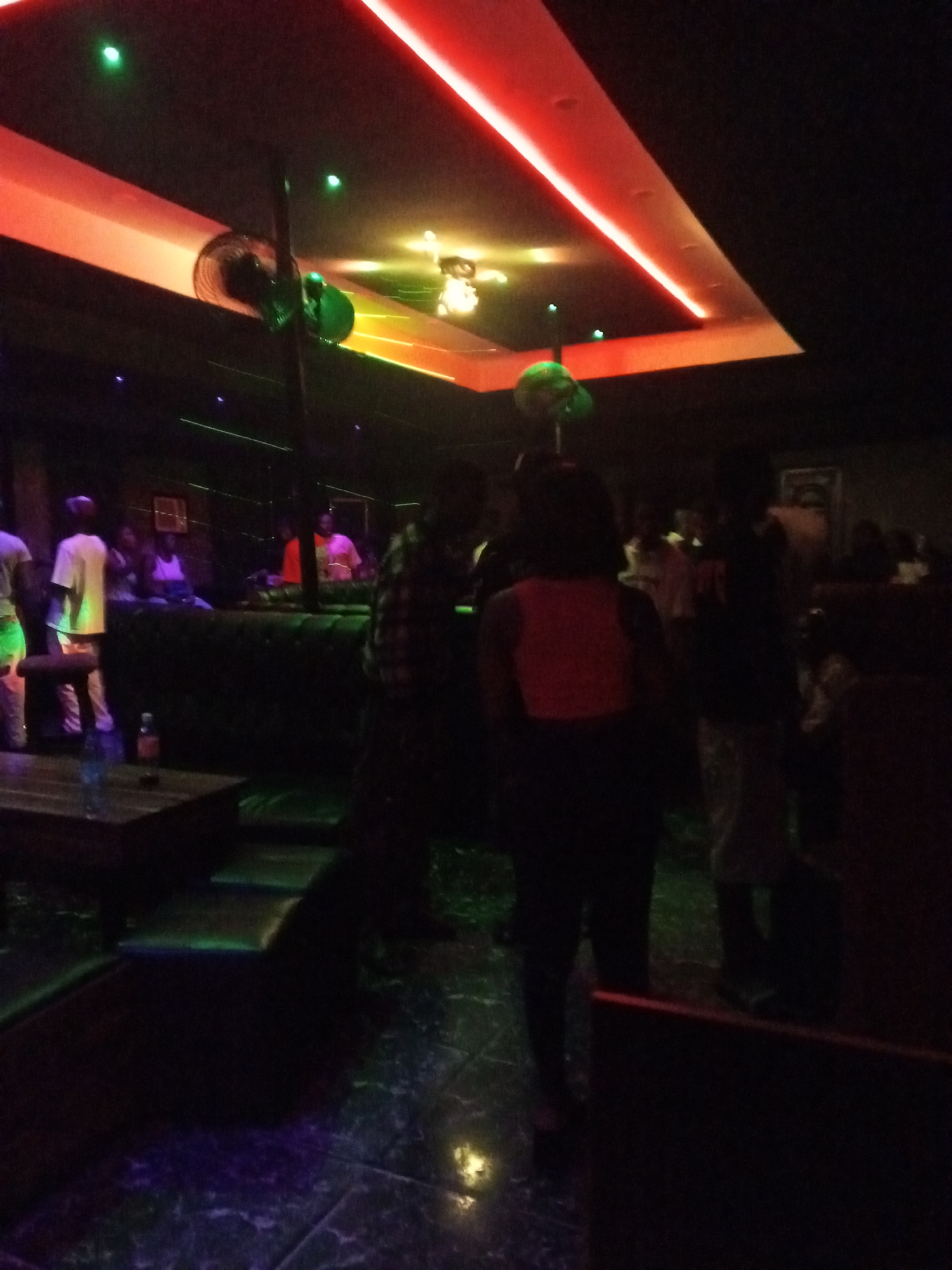 This image depicts the interior of a nightclub or lounge, possibly in Lokoja, Nigeria, though this location cannot be definitively confirmed. Several people are present, some standing and some seated. The lighting is dim with colored neon lights on the ceiling. A ceiling fan and additional decorative lighting fixtures can also be seen. There are tables and upholstered seating areas. The environment appears to be a social gathering or entertainment venue.