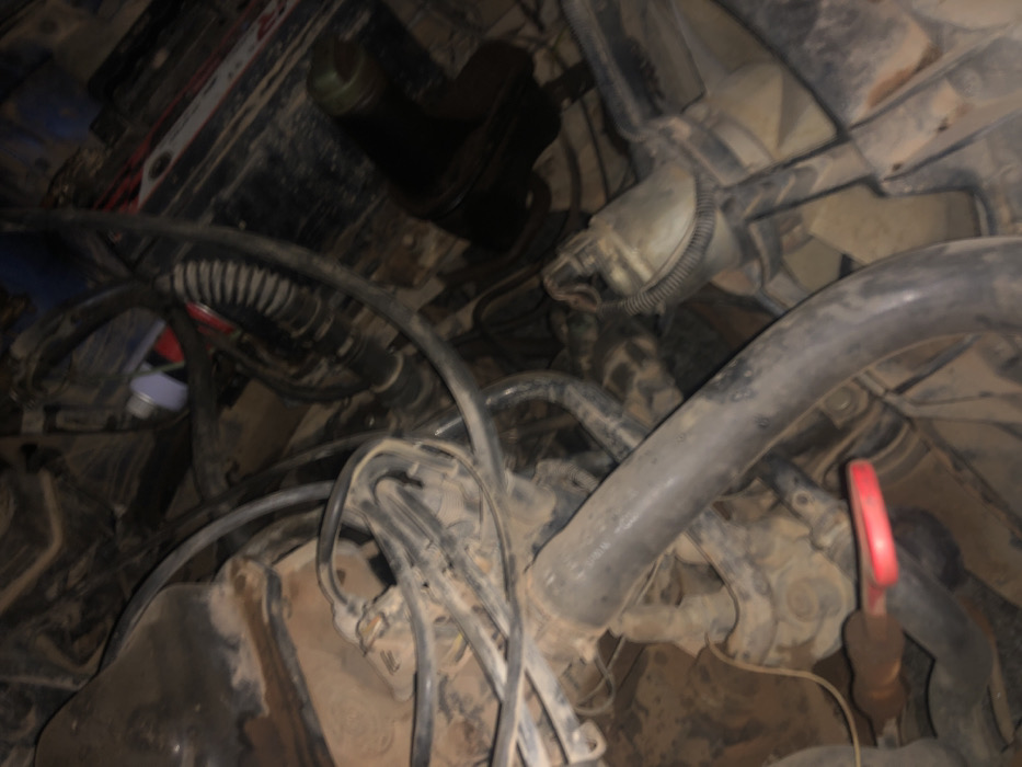 The image shows a dirty engine bay with various hoses and components. A thick, black hose curves across the foreground with a red dipstick nearby. Behind it, numerous smaller black hoses and wires intertwine. A cylindrical, gray component sits partially obscured in the upper-right, while a black, cylindrical object is situated in the upper-center. Overall, the scene is cluttered and suggests an older, unmaintained engine.