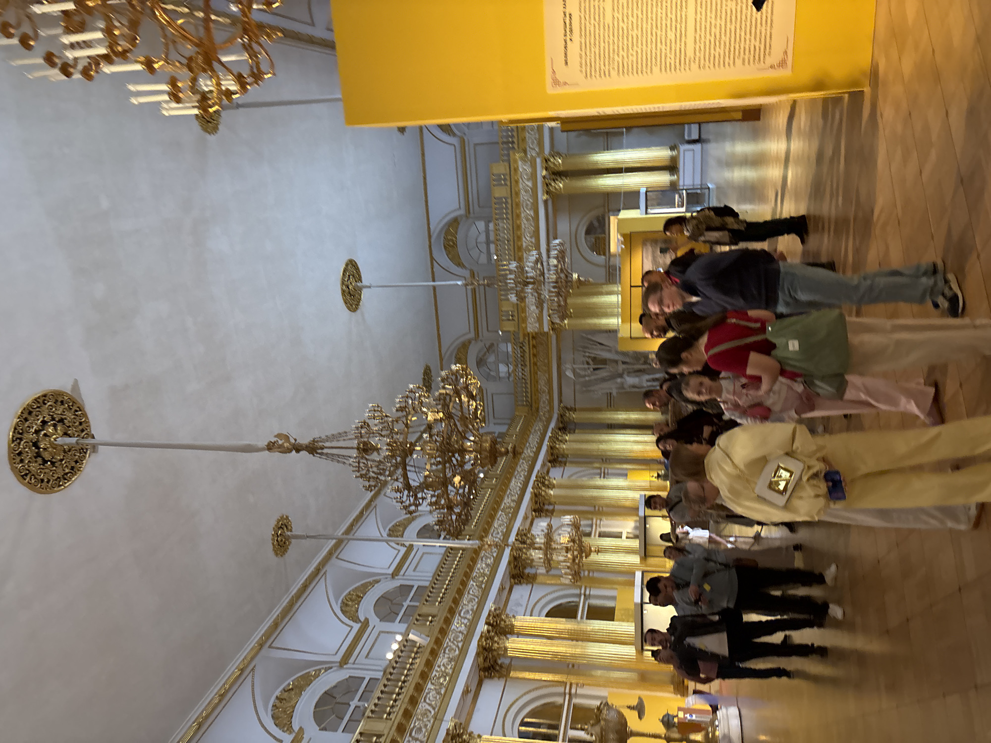 The media file shows an indoor scene with several people standing in a line, seemingly observing something or waiting. The setting appears to be a grand hall, possibly a museum or palace, with ornate gold chandeliers hanging from the ceiling and decorative architectural details like columns and arched windows. The walls are painted a pale color, contrasted by a bright yellow panel at the top, which has some text on it. The floor is made of polished wood.
The people are dressed in casual clothing, suggesting it's a daytime visit. Their postures and expressions are not clearly discernible due to the angle and resolution, but they appear to be engaged with the surroundings. One person in the foreground is wearing a light yellow outfit and carrying a white handbag with a gold clasp. Another person is wearing a blue jacket and grey pants. The overall atmosphere seems calm and orderly.
The lighting suggests it's not nighttime, and there are no indicators of weather conditions as it's an indoor scene. The grand architecture and artistic decorations are prominent features of the setting, pointing towards a location of cultural or historical significance. The text on the yellow panel is mostly illegible in the provided image.
