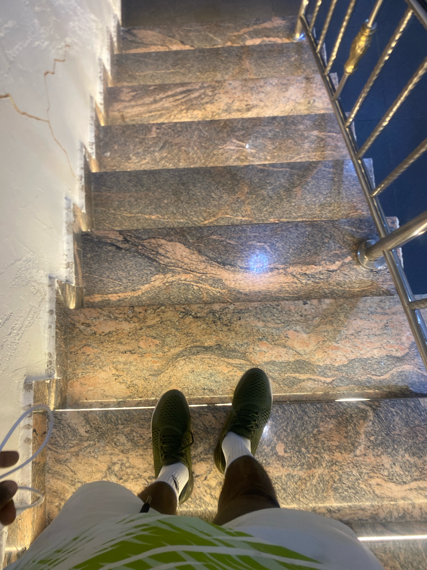 A first-person perspective shot shows a person standing on a marble staircase, possibly in Barden Yana, Nigeria. The person is wearing dark green athletic shoes with white socks, and dark shorts. They are holding a white cord with their left hand. The staircase has a metallic railing on the right side. The walls of the staircase appear to be textured and painted white, with some visible cracking. The marble steps have a distinctive pattern of grey, black, and pinkish-orange veins, and are illuminated from below by embedded lights.