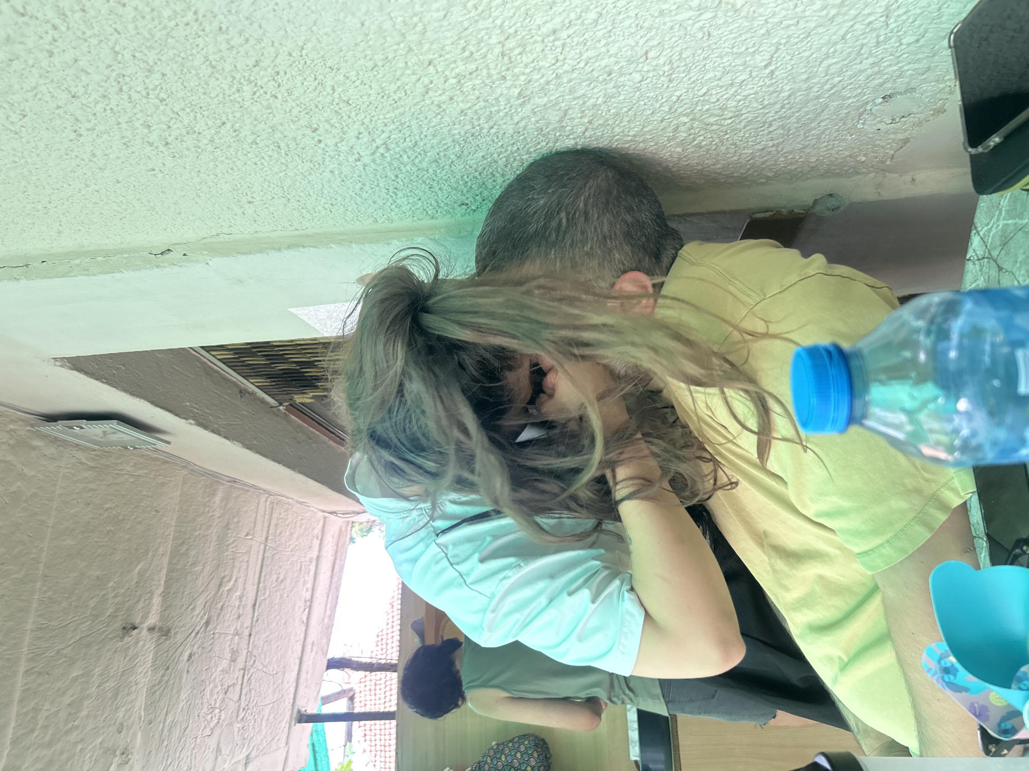 The image depicts an indoor scene, possibly a home or a casual establishment, with two individuals in the foreground. One person, with long, messy brown hair, is leaning over and appears to be kissing or very close to the face of another person, whose head is pressed against a textured, light green wall. The person leaning forward is wearing a light blue t-shirt, and the other person is wearing a yellow t-shirt. In the background, another individual with short dark hair is visible, wearing a green tank top and dark shorts. There are some objects on a table in the foreground, including a blue-capped plastic bottle of water, and what appears to be a smartphone. The walls have a rough, textured finish, and the ceiling is textured and white. The overall environment suggests a casual and possibly rustic setting. The image does not provide any specific visual cues that would place it in Govedare, Bulgaria, or any other specific city.