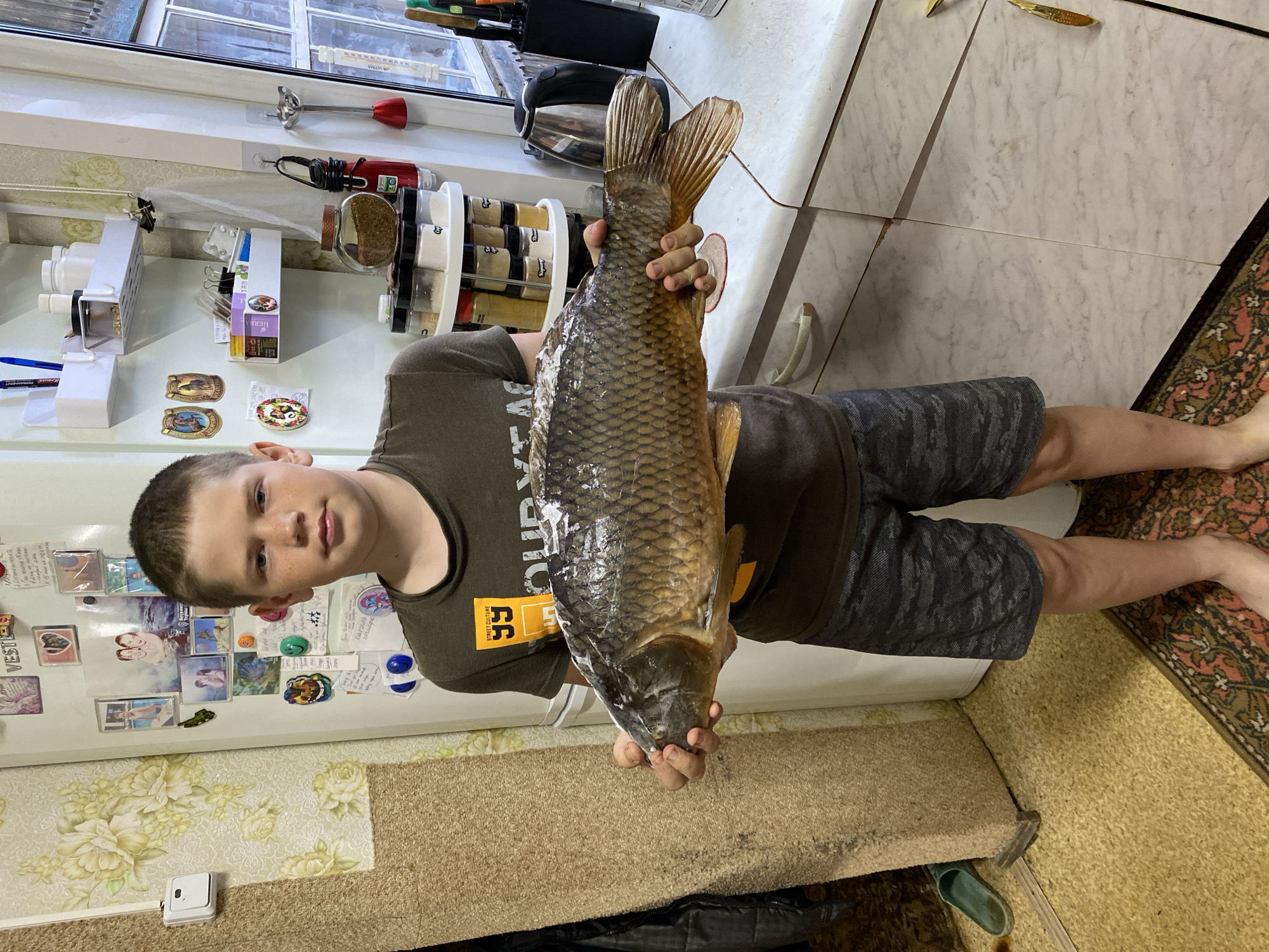 A boy stands indoors, holding a large carp. He is positioned in front of a refrigerator covered with magnets and photos. To his right, there's a tiled countertop with various kitchen items, including a spice rack. The floor has a patterned rug. The location is potentially Баклуши, Russia, based on metadata.
