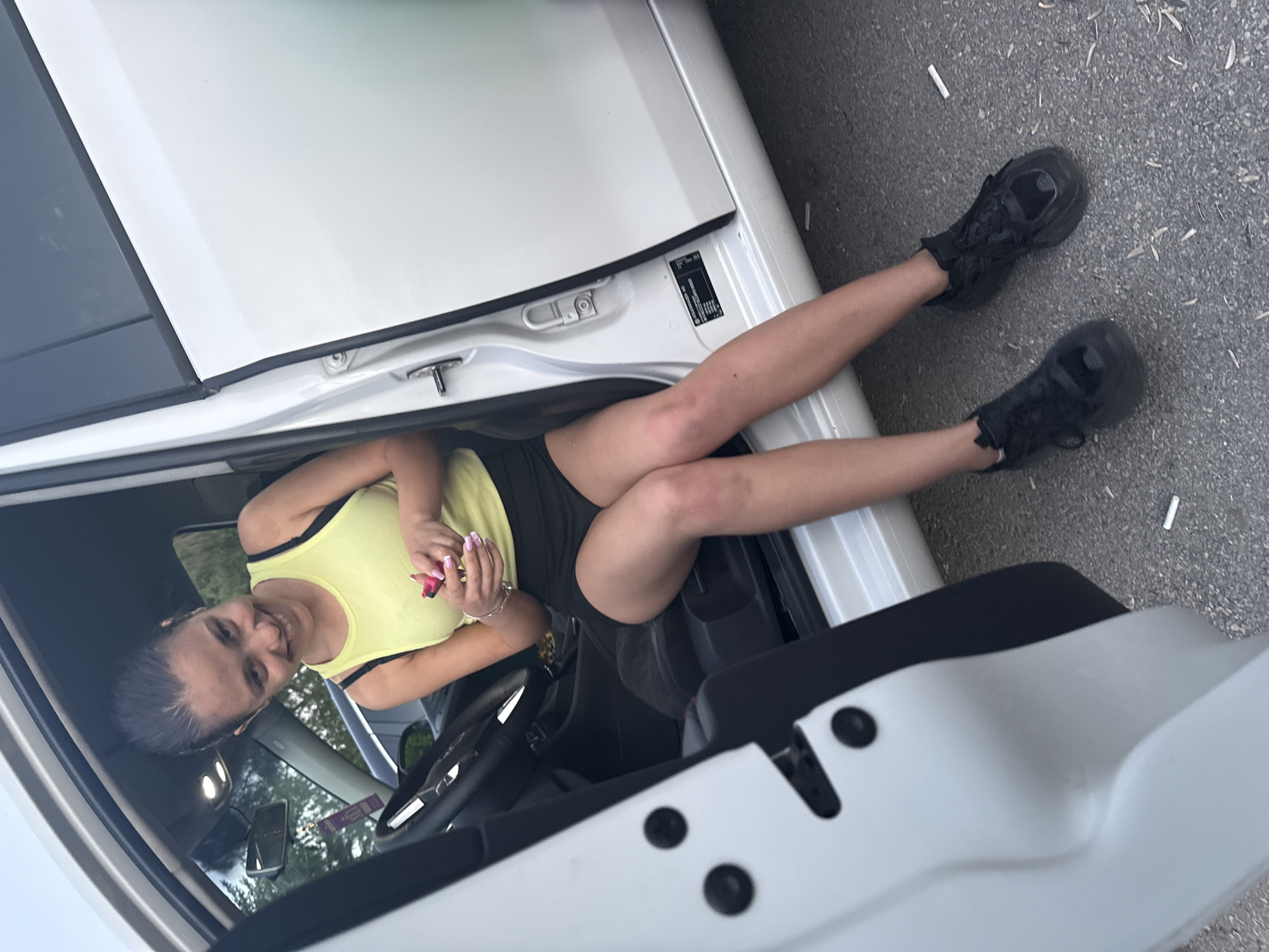 A young woman is sitting in the driver's seat of a white car, her legs extended out of the open driver's side door. She is wearing a yellow tank top with black trim and black shorts. She has dark hair pulled back and is looking at the camera, smiling. She appears to be holding something small and pink in her hands. Her legs are slightly crossed at the knees, and she is wearing black sneakers. The car door is open, and the ground outside the car is asphalt with some small debris. The scene is likely outdoors during the daytime. There is no indication of Pleven, Bulgaria, in the image.