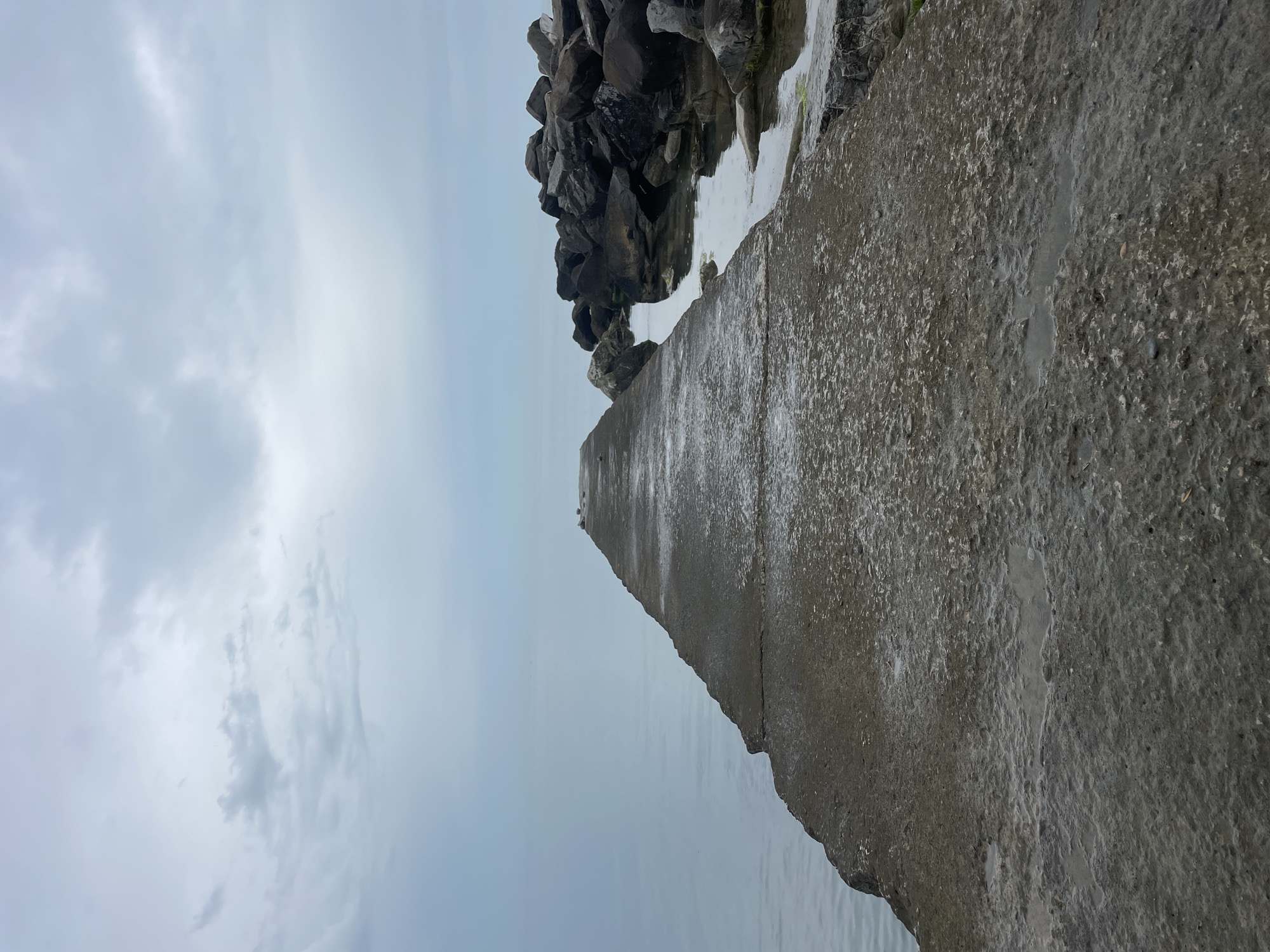The image shows a pier made of concrete, leading out into the water. The pier's surface appears wet, reflecting the overcast sky above. To the side of the pier are large rocks, some partially submerged. The water is calm. The pier is located in the city of Sochi, Russia.