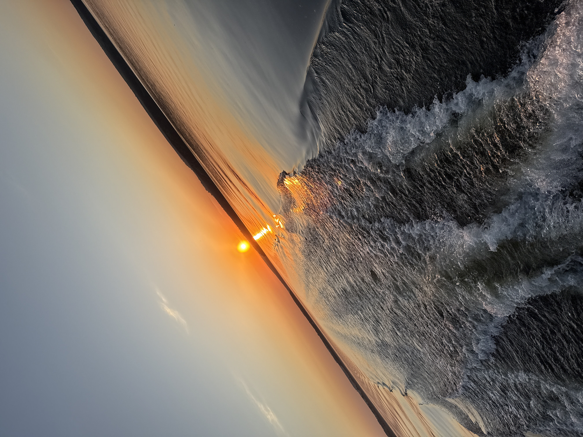 The image shows a sunset over the Votkinsk Reservoir in Votkinsk, Russia. The sun is low on the horizon, casting a golden light across the water. The water is choppy, with waves breaking near the shore. The sky is a mix of gray, orange, and yellow colors. The horizon line is slightly tilted. No people are visible.