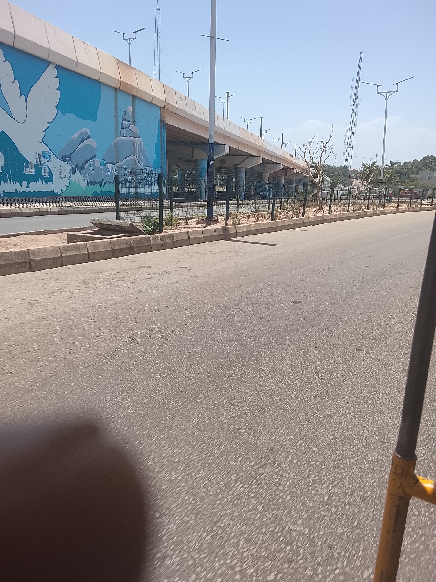 The image shows a section of a road in Jos, Nigeria, with an elevated highway overpass. The overpass is decorated with a mural featuring a white dove and a landscape with rocky formations. A black metal fence runs along the side of the road, separating it from a narrow strip of vegetation. Streetlights are visible along the overpass and beyond. The road surface is gray asphalt. Part of a yellow metal structure is visible in the foreground on the right. The sky is clear and blue.