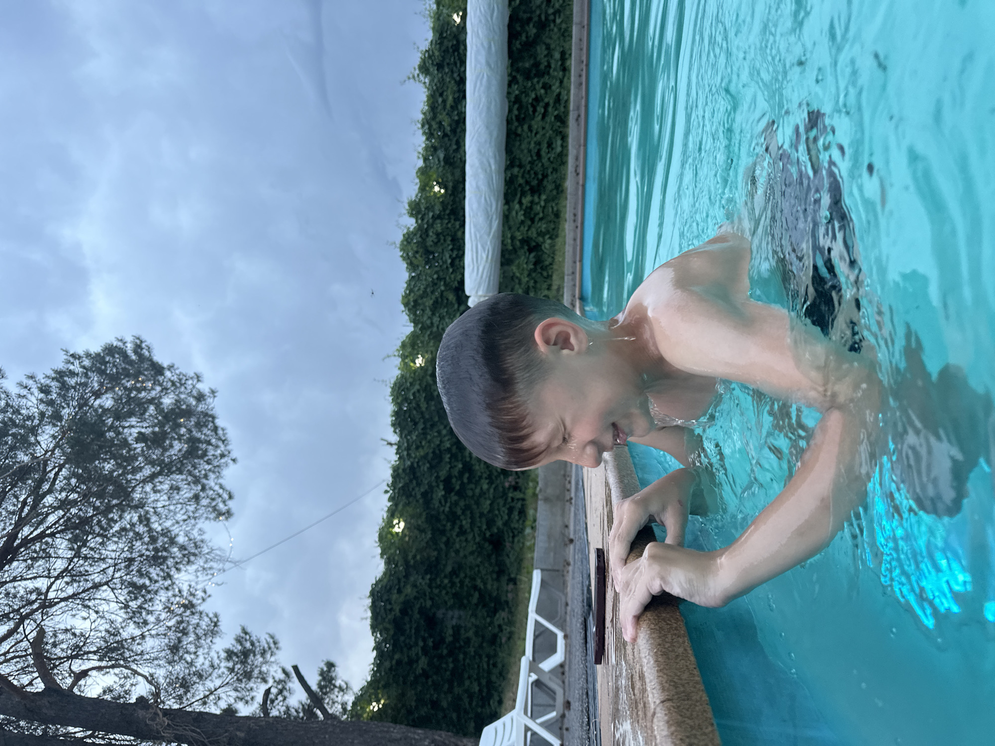 A young person is partially submerged in a swimming pool, gripping the edge with their hands. The pool appears to be outdoors, surrounded by greenery, including a tree and a vine-covered fence or wall. The sky is overcast. White plastic chairs are visible near the pool edge. No information confirming the location as Chaikovsky, Russia is available from the image.