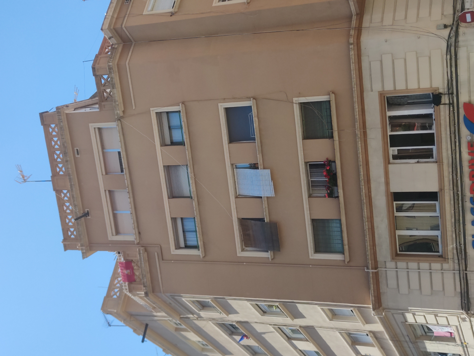 The image shows a multi-story building against a bright blue sky. The building has a light brown facade with rectangular windows, some with visible blinds or curtains. A few windows have flower boxes. The architectural style appears to be European, potentially Mediterranean, given the coloring and detail on the building. The building is likely located in a dense urban area, with other buildings partially visible at the bottom of the frame. Wires and pipes run along the side of the building. A partial sign is visible on the right side, but the text is unclear.