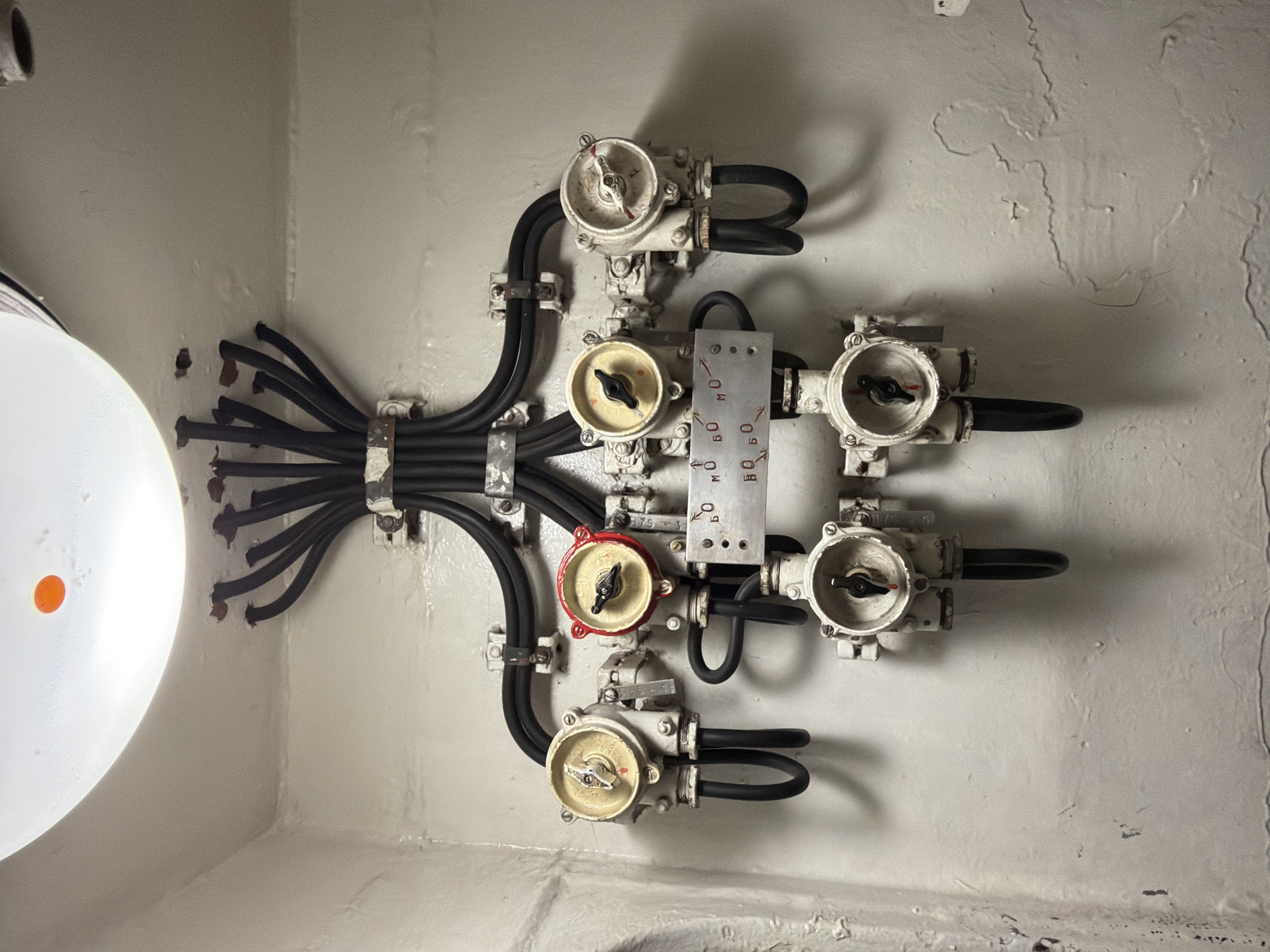 The image shows an electrical panel mounted on a white, textured wall. The panel is comprised of several round, cream-colored junction boxes, each with a rotary switch. One of the switches has a red-painted housing. Black electrical cables snake from a central bundle, connecting to these junction boxes. A metal plate with Cyrillic lettering and numbers is affixed to the panel between some of the boxes. A circular light fixture with an orange spot is visible on the ceiling. The environment appears to be an interior space, possibly utilitarian or industrial, characterized by the unadorned wall and exposed wiring. The scene does not contain any identifiable people. The image is not identifiable as being located in Murmansk, Russia.