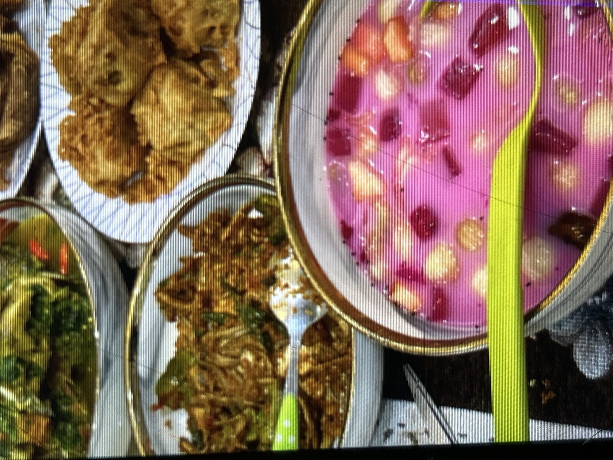 The image displays an overhead view of a table setting featuring several dishes of Indonesian food. In the foreground, a bowl of pink dessert with diced fruits like strawberries and possibly lychee is visible. A bright green spoon is partially submerged in the dessert. Behind this, to the left, is a bowl filled with stir-fried noodles or rice with vegetables and what appears to be shredded meat or fish. Further back and to the right, a plate holds several pieces of fried fritters, possibly tempeh or tofu based. The dishes are arranged on a textured, dark surface, likely a tablecloth. The lighting suggests an indoor setting. The image does not provide any specific information about the location in the city of Depok, Indonesia, or any people present.