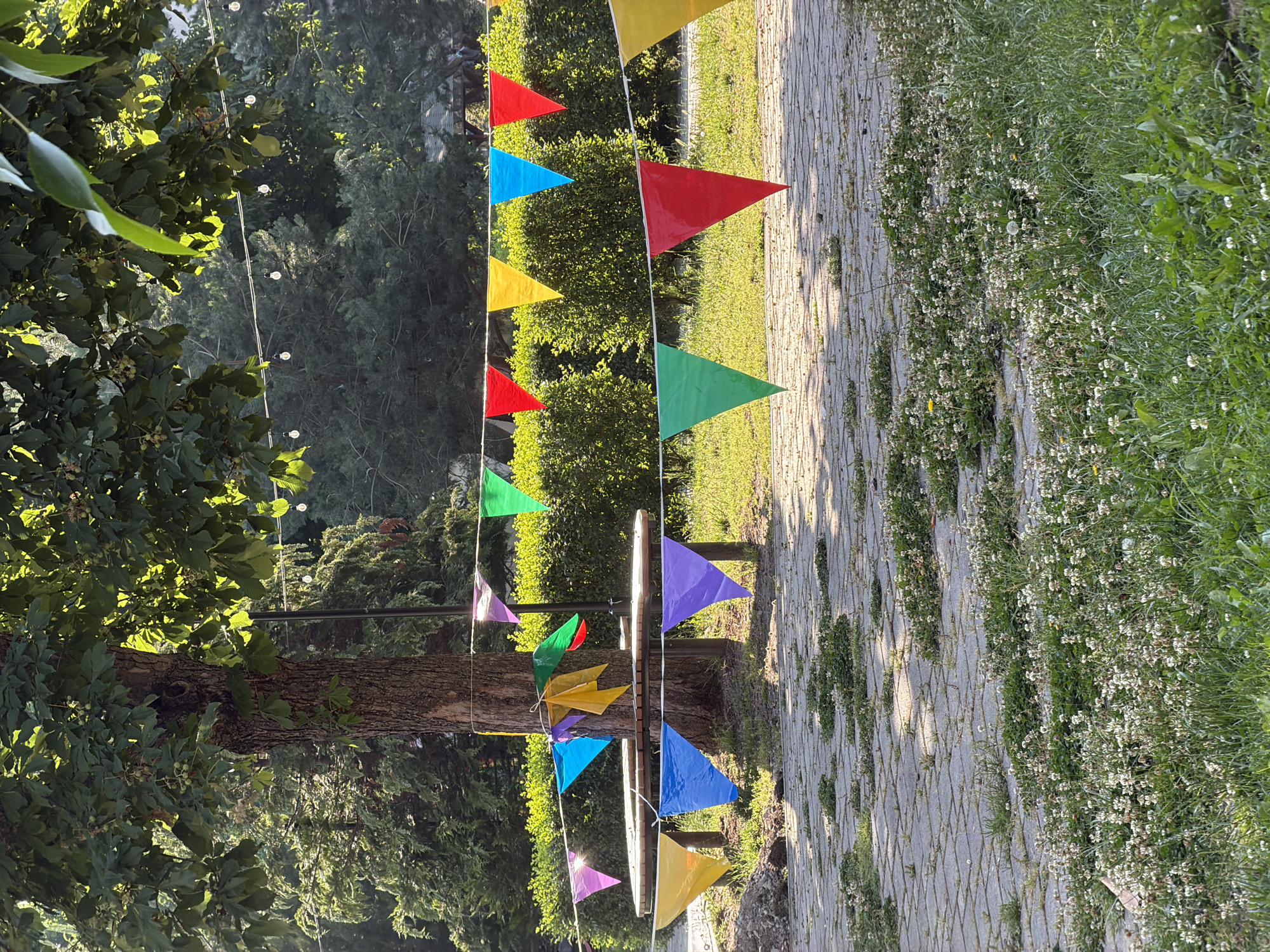 A festive outdoor scene is depicted in a city park. A large tree stands prominently in the mid-ground, its leaves dappled with sunlight. Strung between trees and poles are lines of colorful triangular flags in shades of red, yellow, purple, green, and blue, creating a festive atmosphere. A circular wooden picnic table is situated near the tree, with more flags draped around its legs and on the table itself. In the background, dense green hedges form a backdrop, interspersed with more trees. String lights are visible in the upper portion of the image, stretching across the scene. The ground in the foreground is paved with large gray interlocking stones, with patches of grass and small white wildflowers growing in between. The overall impression is a cheerful and inviting outdoor gathering space. The location is identified as a city park in Sofia, Bulgaria.