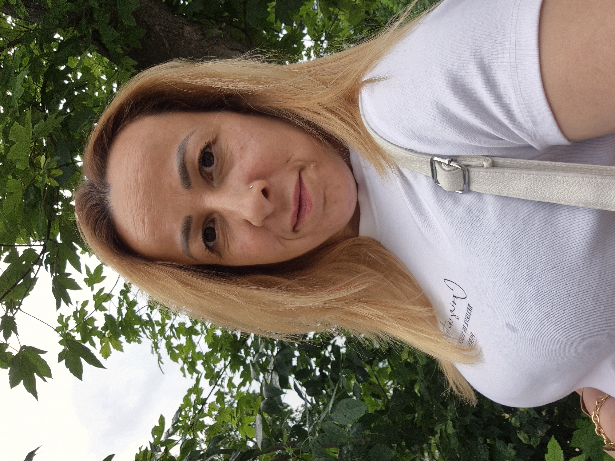 A frontal, close-up photograph features a female subject whose shoulders and head are framed by natural outdoor elements. She has light blonde hair, parted off-center to her left, with darker roots visible at the scalp, falling in a straight or softly wavy style to shoulder length. Her face is oval, displaying visible forehead wrinkles and fine lines around her dark brown eyes, which are directed toward the camera. Her eyebrows are dark, well-defined, and appear groomed. A small, silver-toned stud piercing is present on her left nostril (viewer's right). Her lips are closed, exhibiting a faint, neutral expression or slight smile. Her skin tone is fair, with a few small moles or freckles visible on her cheeks.
She is wearing a white, short-sleeved, ribbed t-shirt. Reversed black text is visible on the chest area, appearing to read "BETTER IN YOUR" and "PARIS" below it. A light-colored (off-white or cream) shoulder bag strap, equipped with a rectangular silver buckle, crosses over her right shoulder (viewer's left). A gold-colored, chunky link chain bracelet is partially visible on her left wrist (viewer's right).
The background consists of dense green foliage from deciduous trees, some with visible darker trunks, suggesting an outdoor, possibly park-like, setting in the city of Unknown, Unknown. The sky, visible through the leaves in the upper left, is overcast and light gray, indicating daylight conditions without direct sunlight. No direct actions or interactions beyond posing for the photograph are discernible.