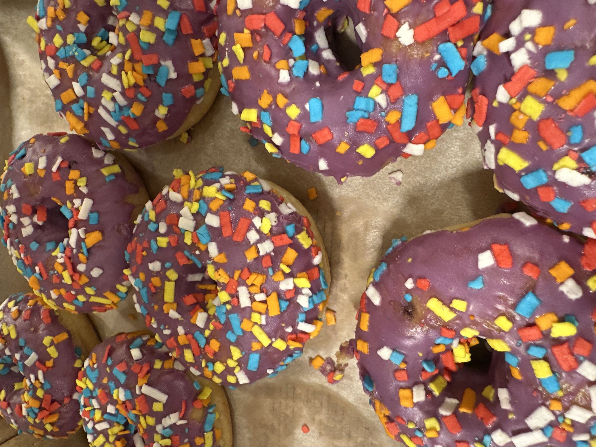 The image shows a collection of glazed donuts with colorful sprinkles arranged in a container lined with parchment paper. The donuts are frosted with a purple glaze and covered with rectangular sprinkles in shades of white, blue, yellow, and orange. The location is unknown and there are no people visible in the image.