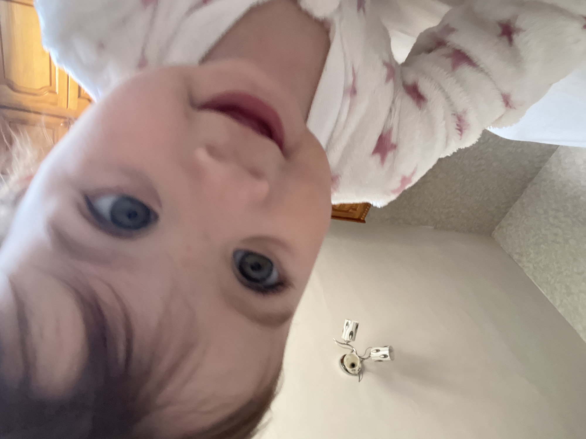 A low-angle, close-up image captures a child's face, likely a baby or toddler, wearing a white robe with pink stars. The child has fair skin, blue eyes, and brown hair. Behind the child, a wooden cabinet is partially visible on the left. To the right and below the child, the ceiling is visible with a chrome light fixture mounted on it. The background includes textured white walls. There is no information in the image to establish that the location is Elena, Bulgaria.