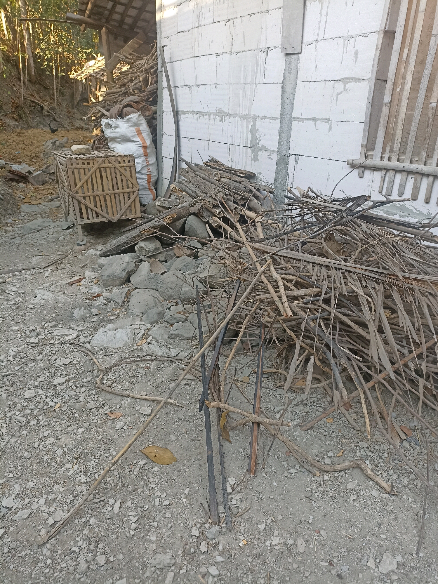 A construction site in Widoro, Indonesia, shows the exterior of a white brick building under construction. A pile of dried branches and palm fronds is located in front of the building. To the left, a wooden cage and a large white bag filled with material are visible. The ground is covered with gravel and scattered rocks. Trees are visible in the background.
