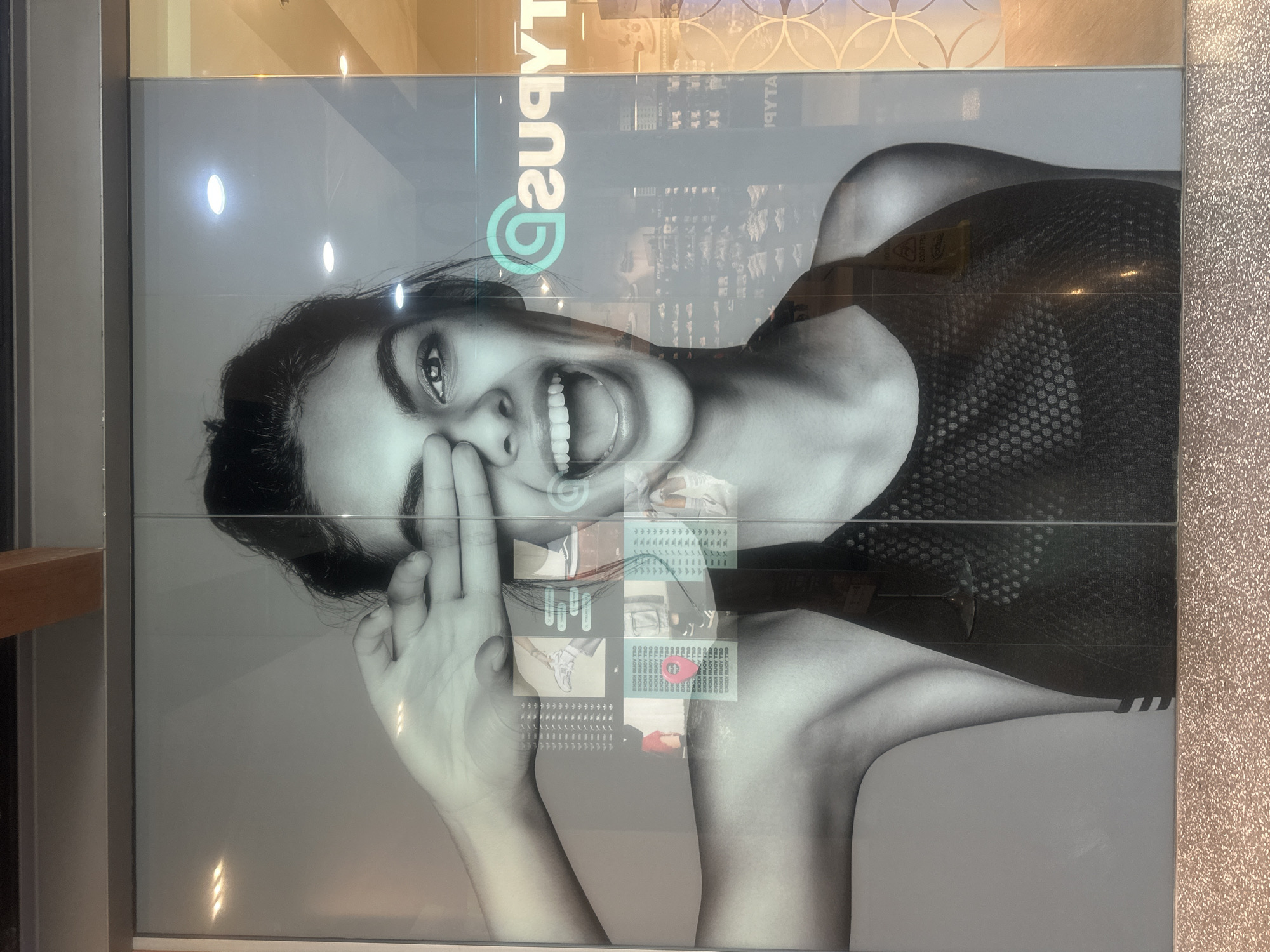 A large black and white advertisement is displayed on a glass storefront. The advertisement features a close-up of a young woman's face and upper body, with her right hand covering her eye and an excited expression. She appears to be wearing a sports bra. Scattered across the image are smaller, superimposed images of athletic shoes and clothing, as well as textual elements in white and turquoise, including a logo that reads "asvtyus". The storefront is located in Coffs Harbour, Australia, and is illuminated by interior lighting, with some reflections visible on the glass. The foreground of the image shows a speckled, light-colored surface, possibly a floor or a base to the display.