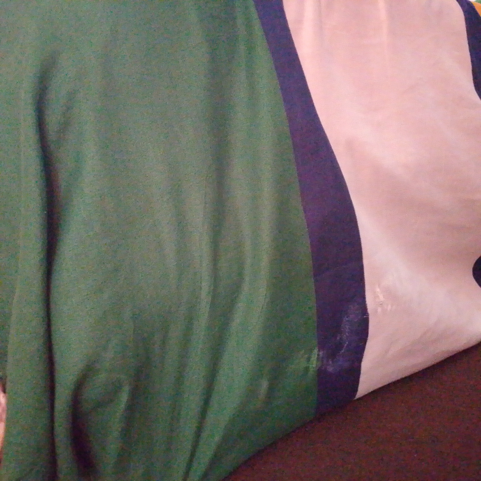 The image shows a close-up view of a fabric. The fabric is predominantly green and appears to have vertical folds. A dark vertical stripe runs down the right side of the green fabric, adjacent to a lighter colored fabric, possibly pink or light peach, which also exhibits folds. The lighting is somewhat dim, creating shadows that accentuate the fabric's texture and folds. There is no discernible background or environmental context that would allow for identification of a specific location in Jalingo, Nigeria. The image focuses solely on the patterned textile.