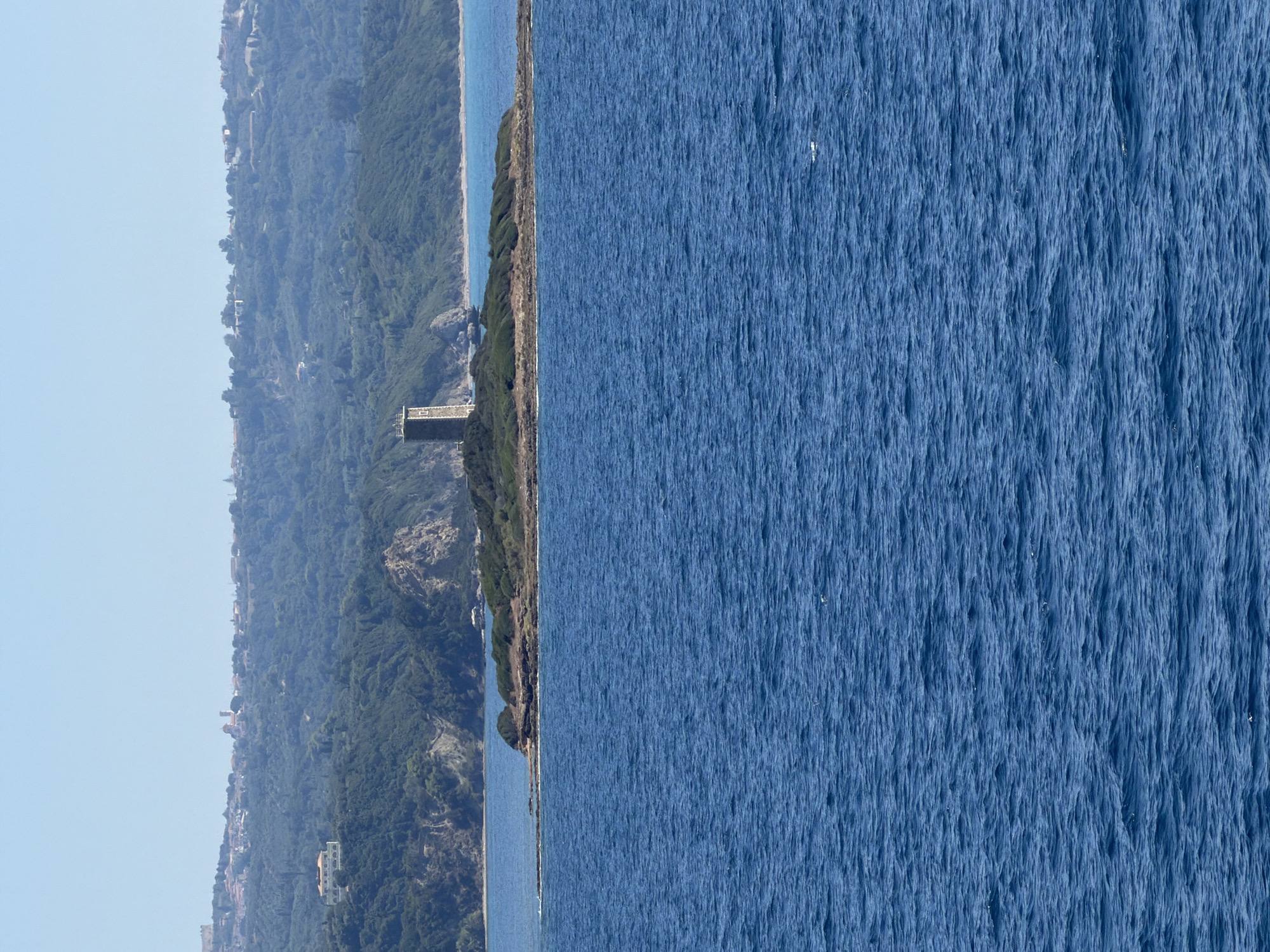 A stone tower stands on a small, rocky island covered in green shrubbery. The island is surrounded by calm, blue water. In the background, a hilly, forested coastline is visible with a scattering of buildings and structures, including what appears to be a settlement or village along the ridge. The sky is clear and pale blue. This scene is the Kastro of Kyllini, located in the Municipal Unit of Kastro - Kyllini, Greece.