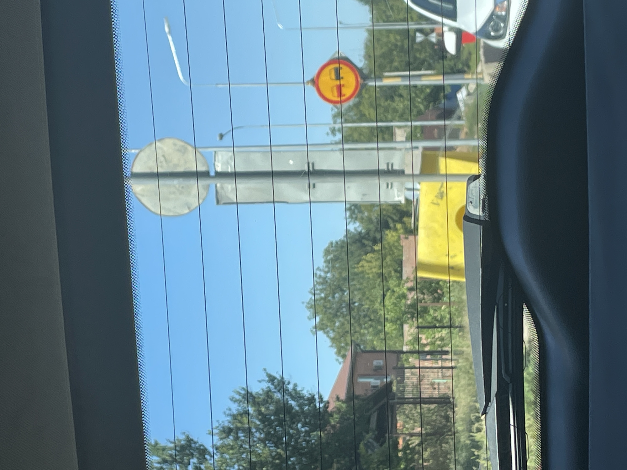 A view through a car's rear windshield shows a street scene in Rostov-on-Don, Russia. The windshield has horizontal defroster lines. In the background, there are trees, buildings, and a large yellow structure. Traffic signs are visible, including one that appears to be a "no passing" sign. A white car is parked on the right side of the frame. The sky is clear and blue. The overall scene suggests a sunny day.